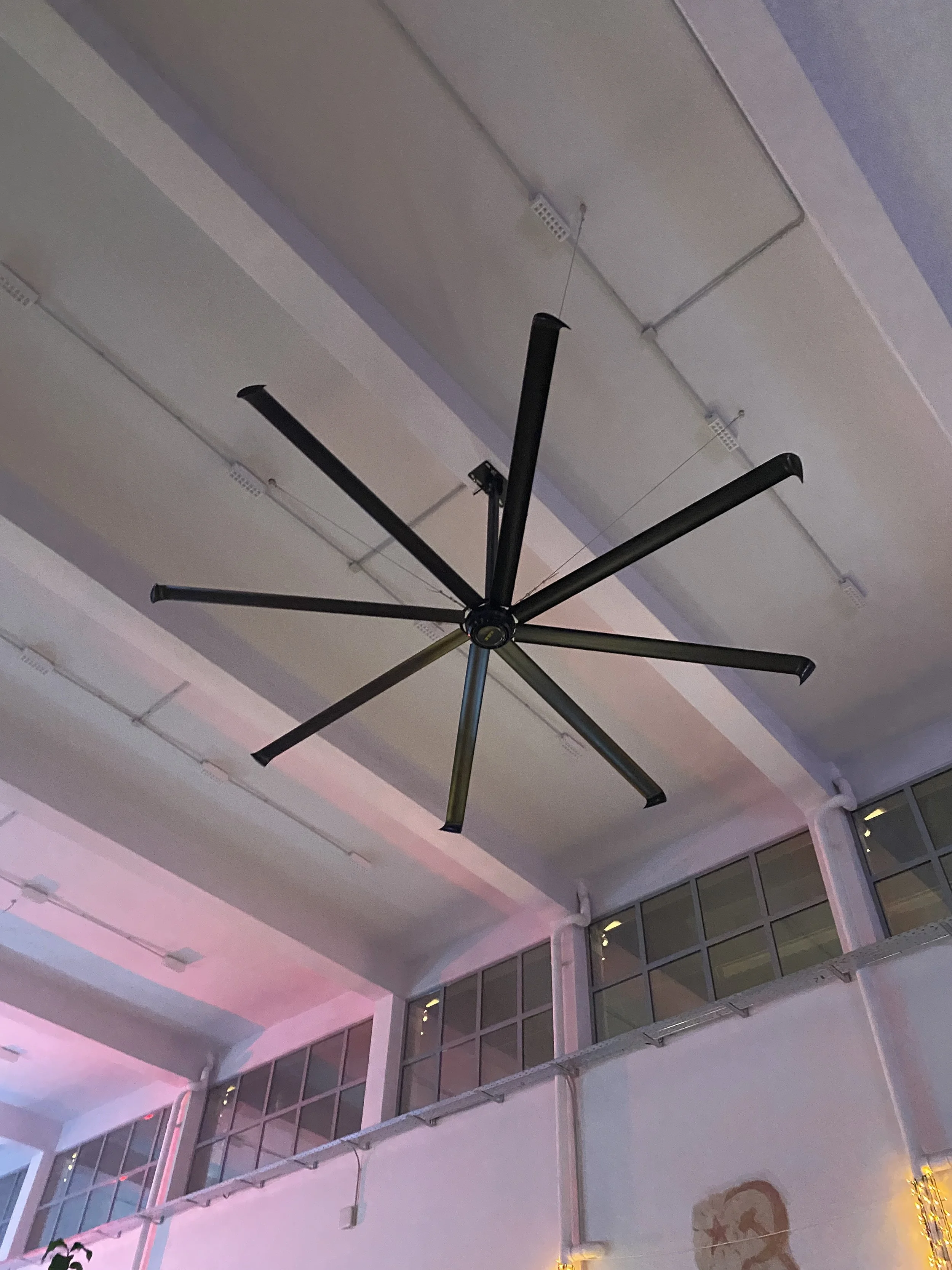 Ceiling with a hanging black fan and visible electrical wiring and ventilation ducts, with large windows and some pink and yellow decorative lighting.