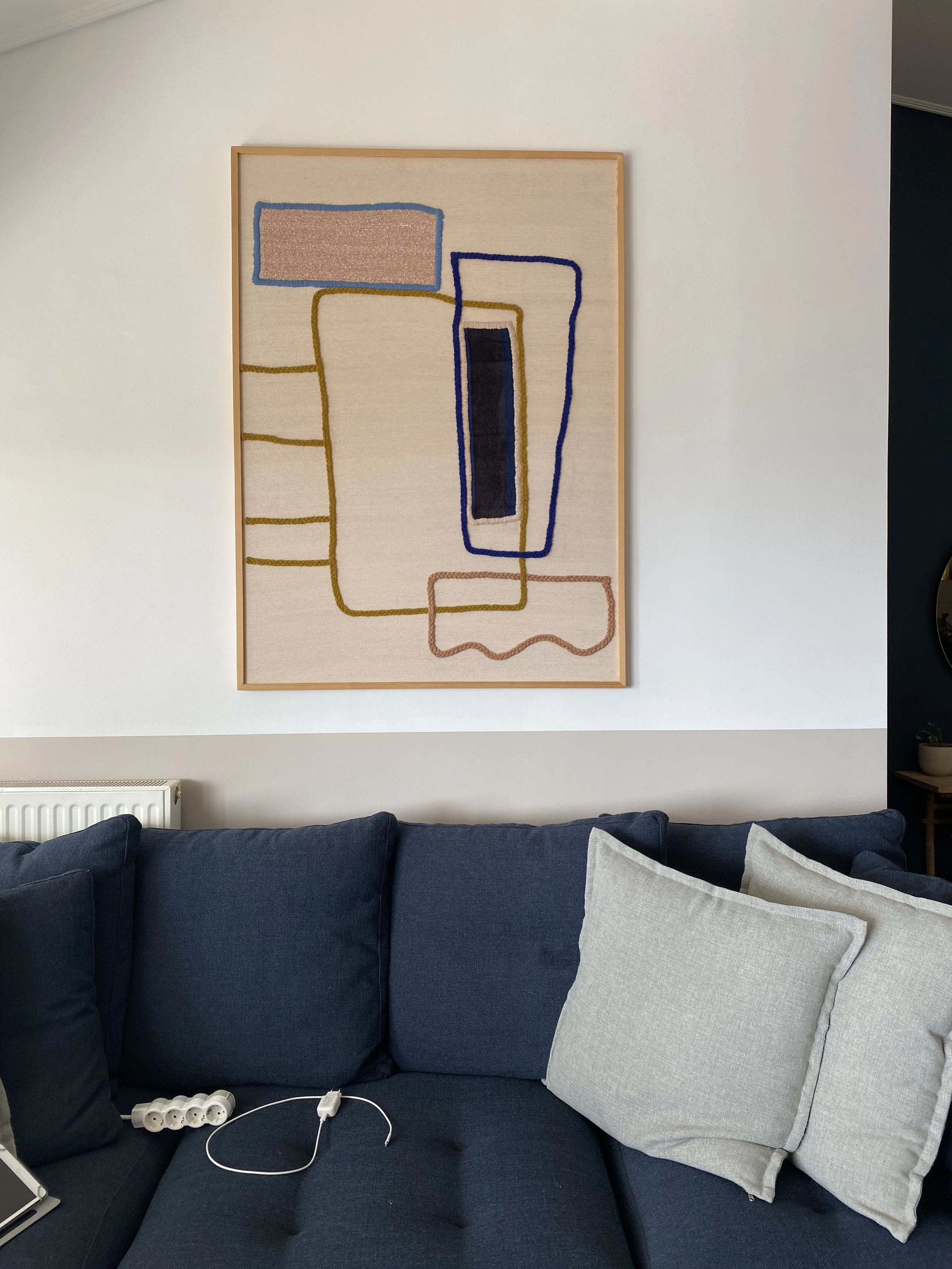 Living room with a dark blue couch, light gray pillows, a wall art piece with abstract line art, and a white power strip with cords on the couch.