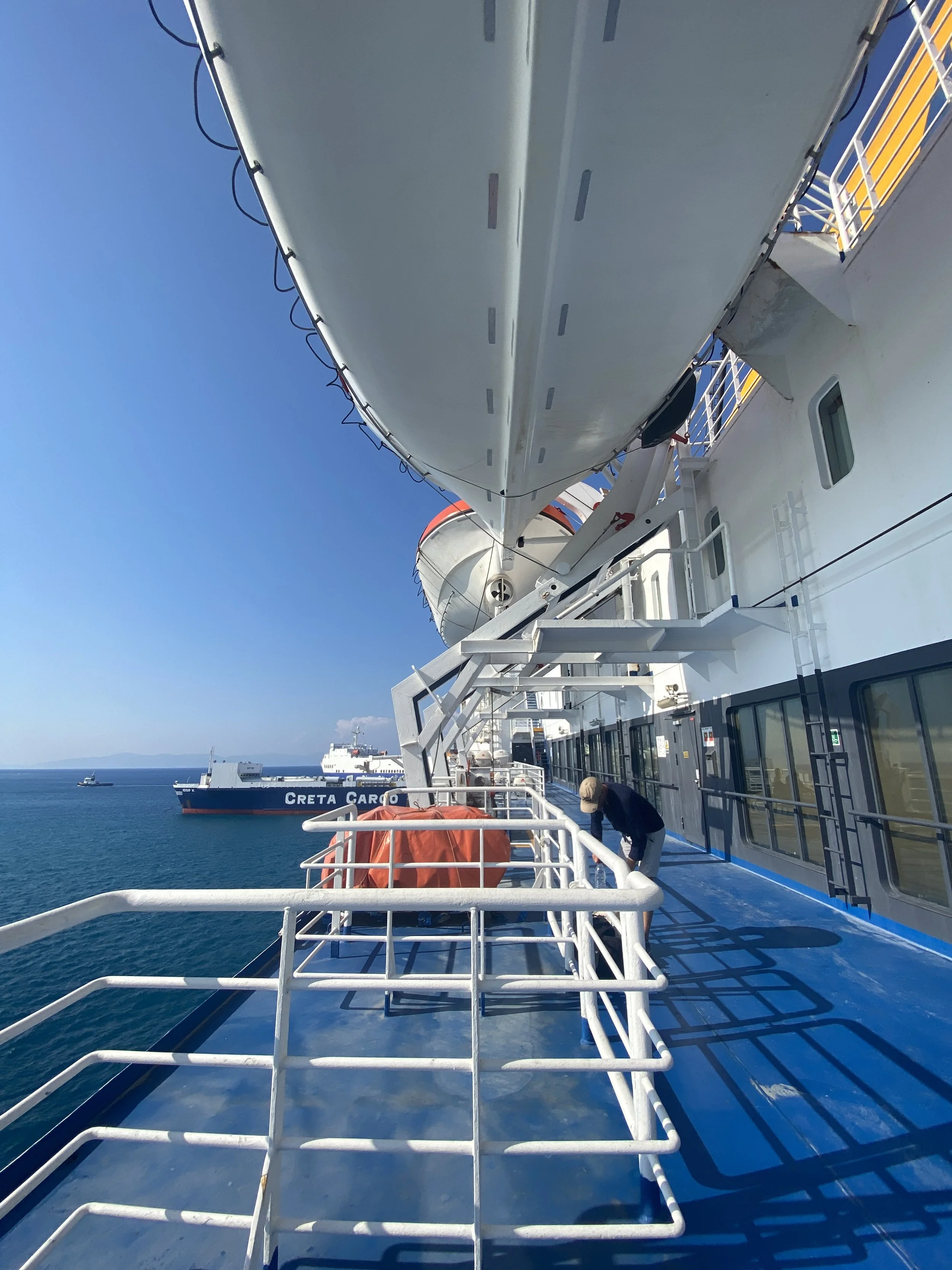 View of the side of a cruise ship with a person cleaning the deck, cargo ships and ocean in the background, and clear blue sky.