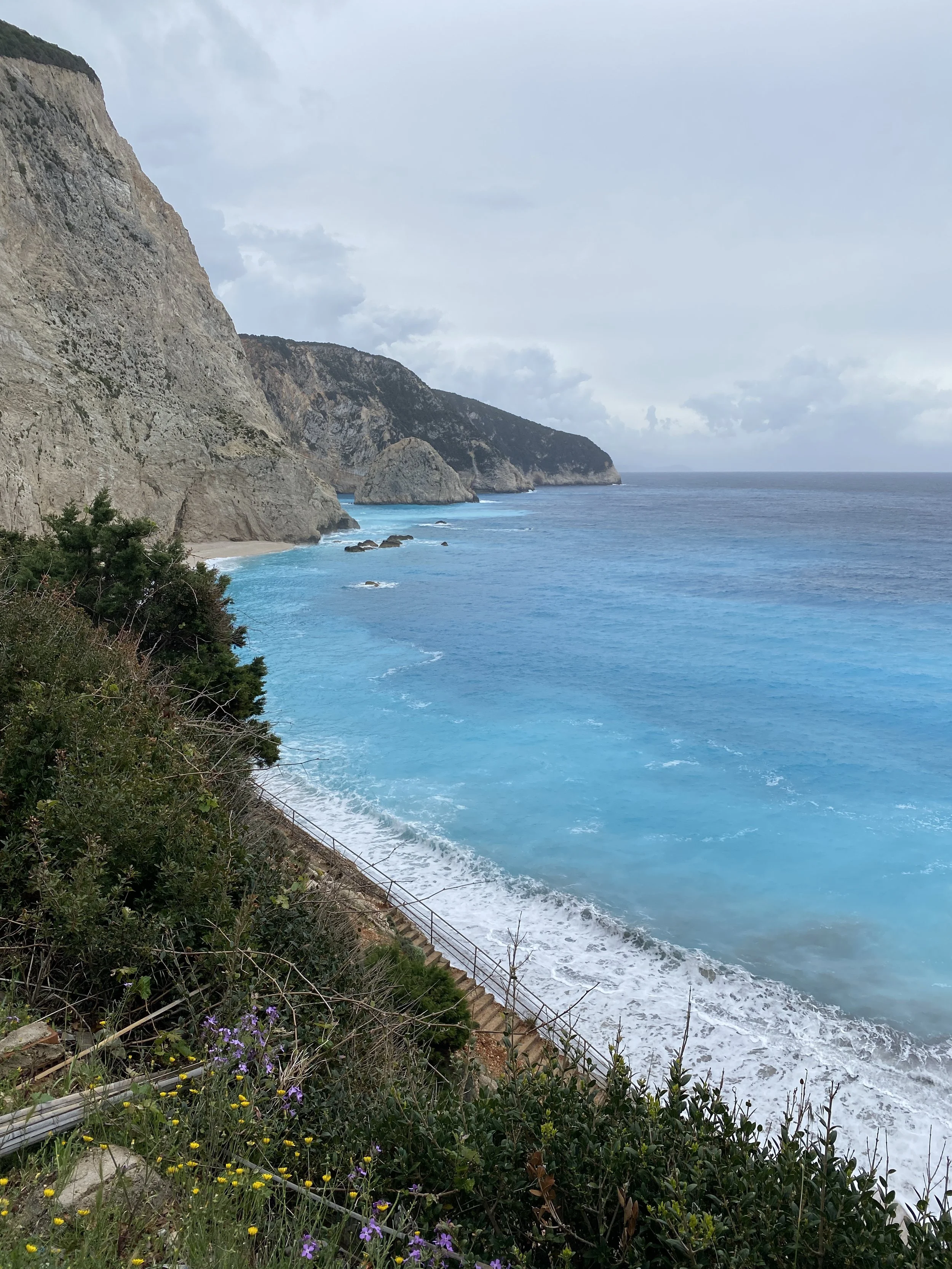 A rugged coastline with steep cliffs, a staircase leading down to a pebbled beach, and the ocean with turquoise waters.