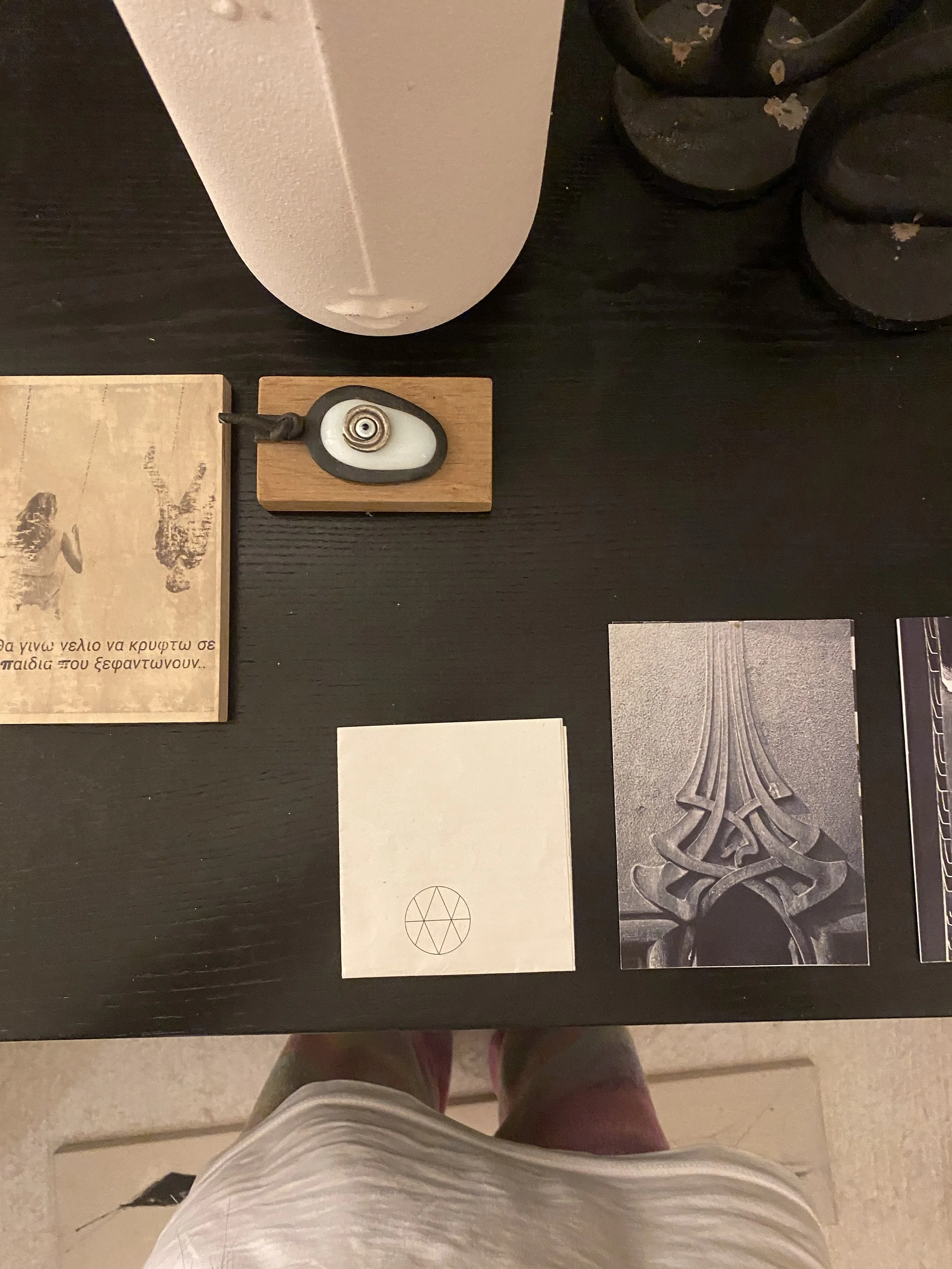 A black table with a white speaker, a wooden object with a black and white stone, a small card with a geometric symbol, and a black decorative item. A person's white shirt and pink pants are visible at the bottom.