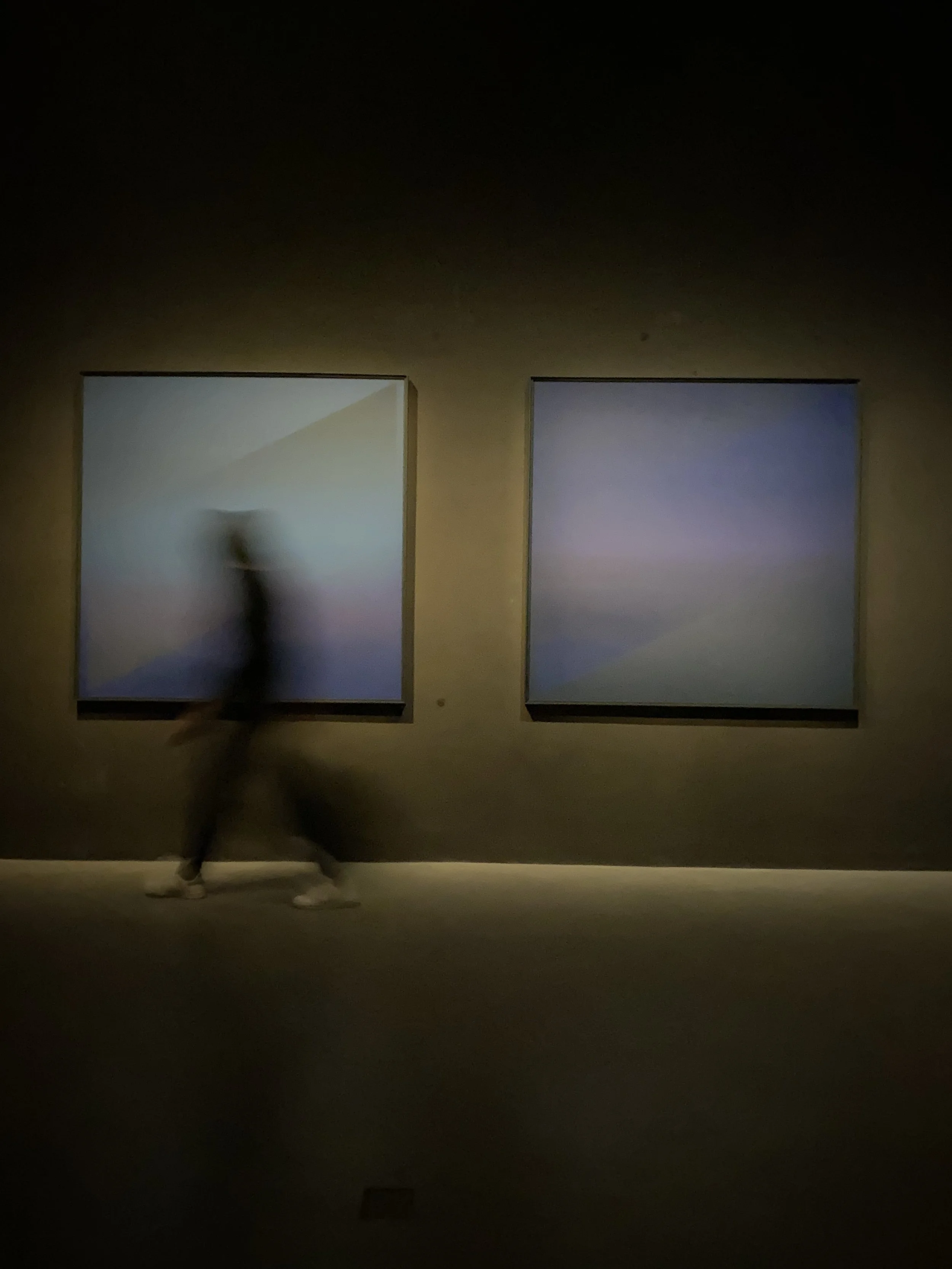 A blurred person walking past two abstract art paintings in a dimly lit art gallery.