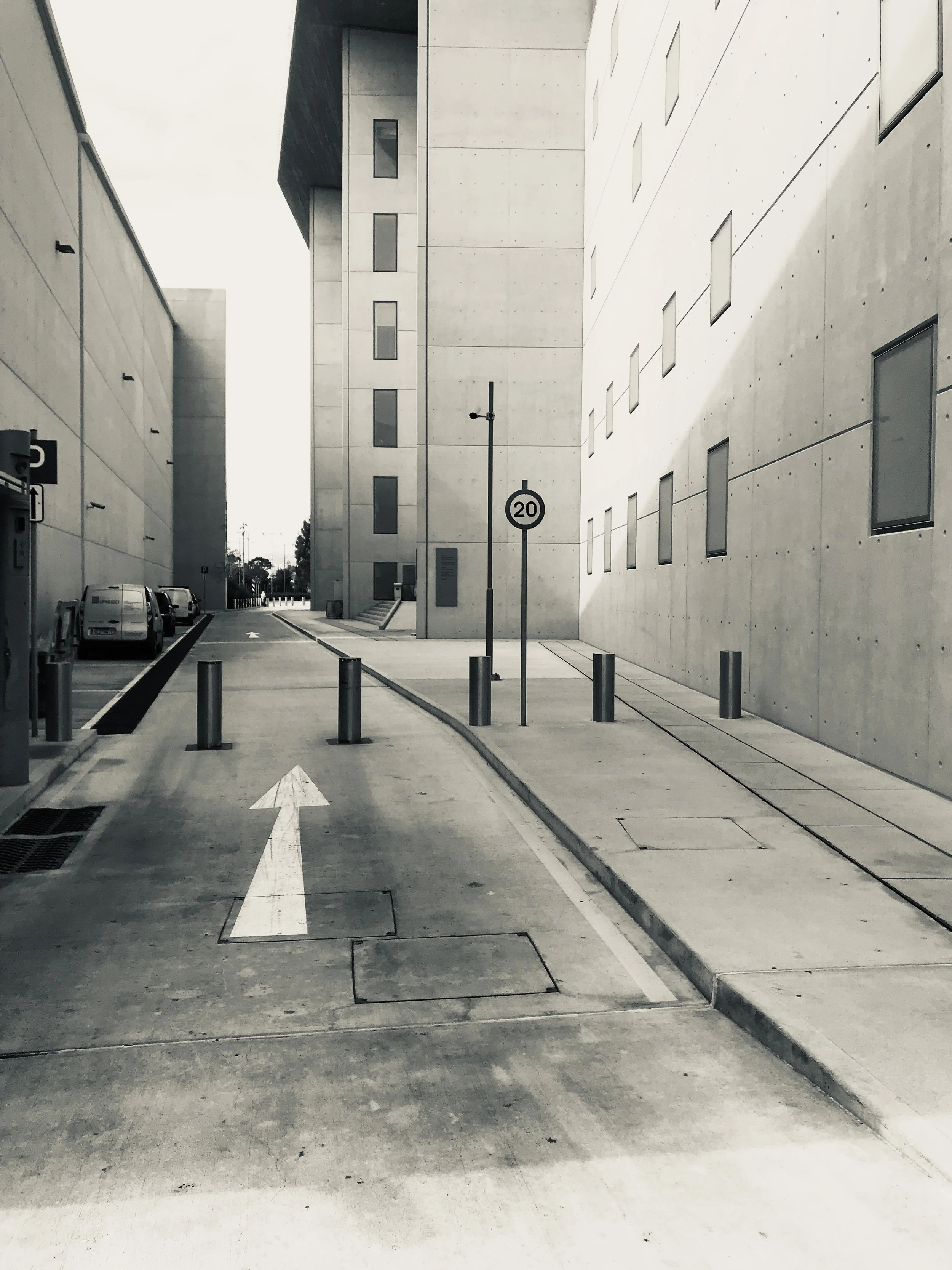 An urban alleyway with modern concrete buildings on either side. There are parking spots with parked cars on the left, a 20 mph speed limit sign, and a one-way arrow on the ground pointing forward. Several short bollards are along the sidewalk and st