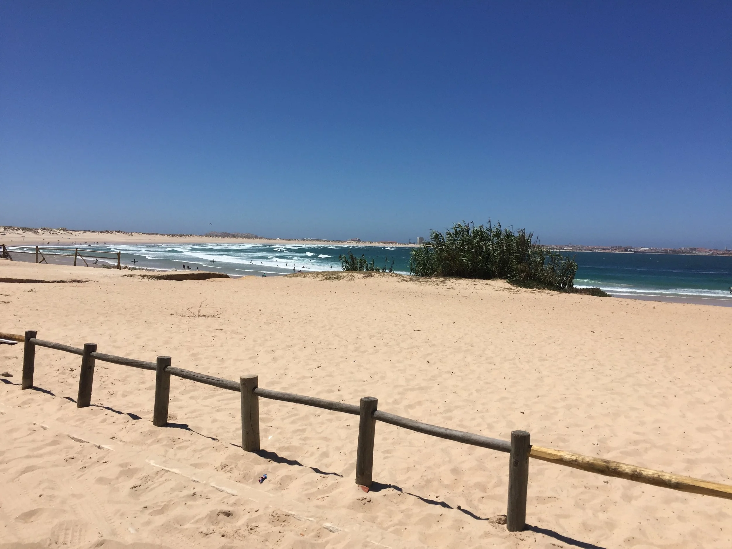 A sandy beach with a wooden fence in the foreground, the ocean with waves in the middle ground, and a clear blue sky with a distant shoreline and some buildings in the background.