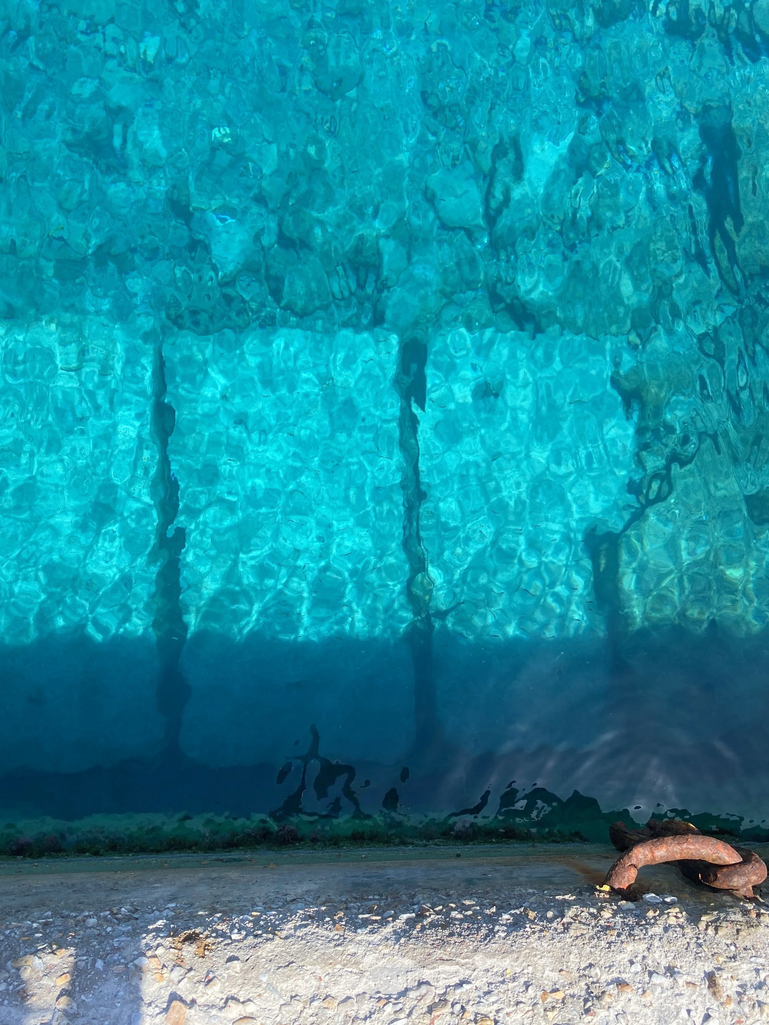 Photo taken from above of a swimming pool with clear blue water, showing the pool's tile floor and a rusted metal pipe at the water's edge.