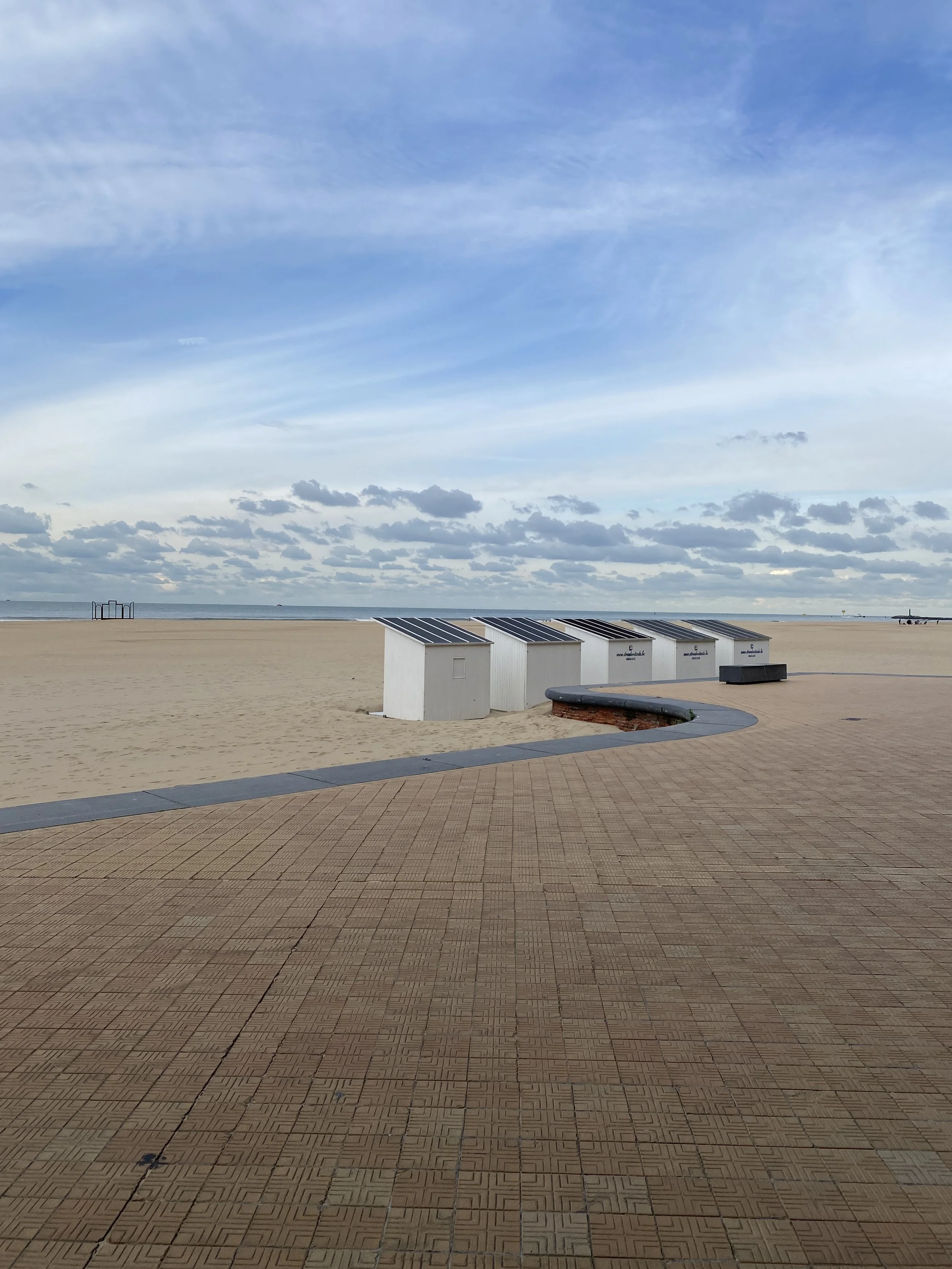 Empty beach with small white beach huts that have solar panels on their roofs, a paved walkway, and a cloudy sky.
