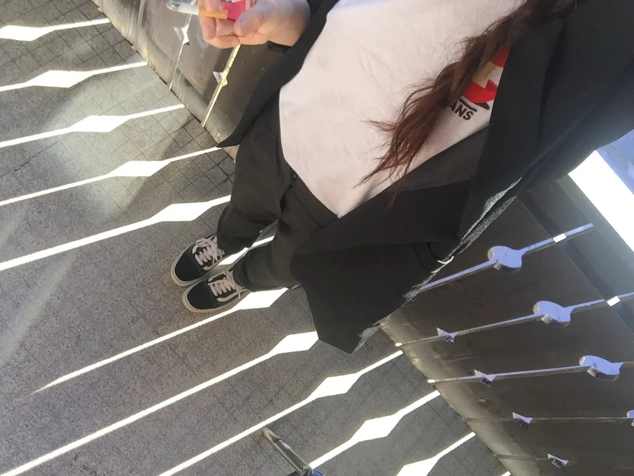 Person standing on a balcony with black railing, wearing sneakers, black pants, a light-colored T-shirt, and a black jacket, holding a lit cigarette.