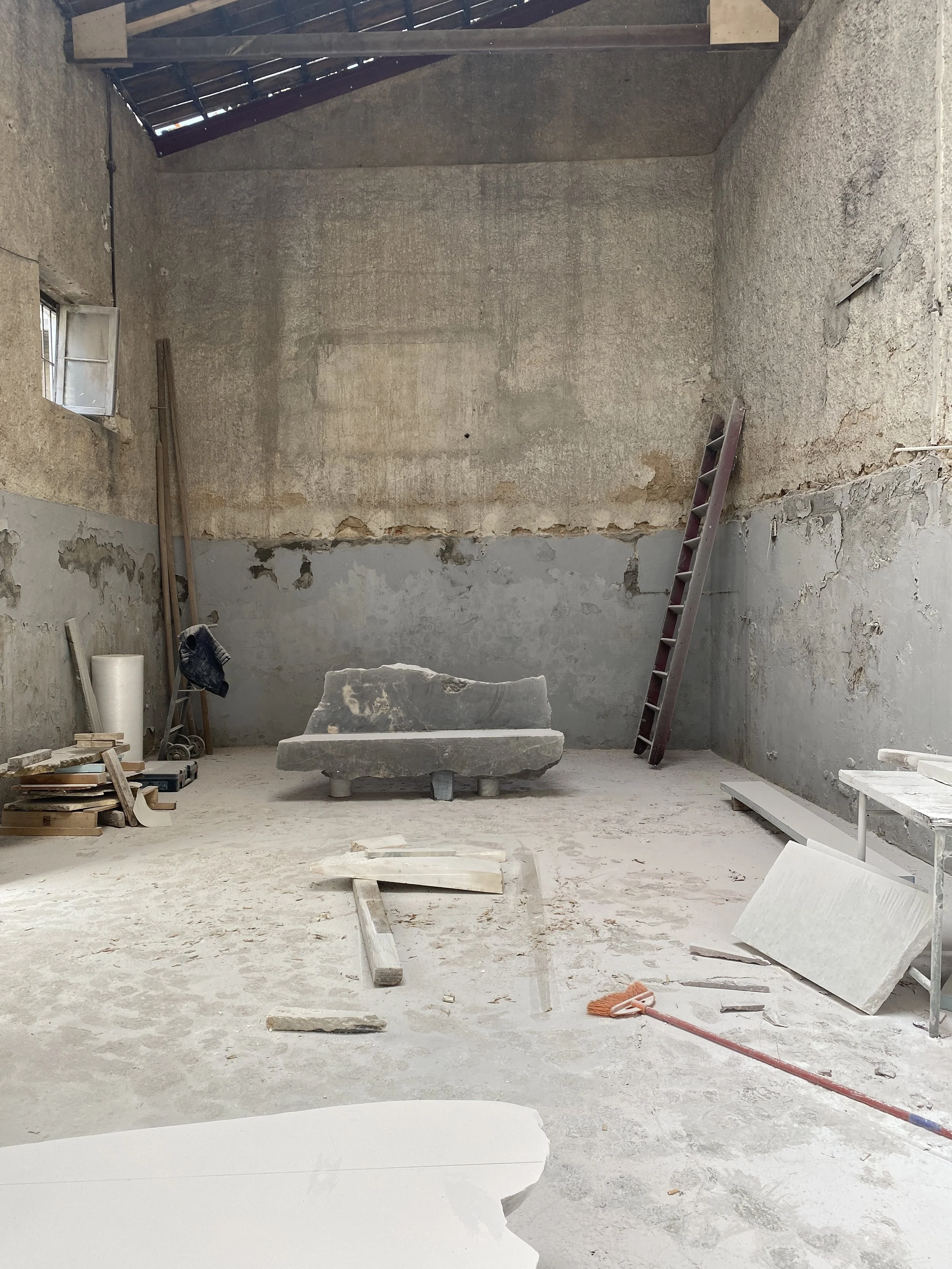 Interior of an unfinished room under construction with a concrete bench, ladder, tools, and construction materials scattered on the dusty floor.