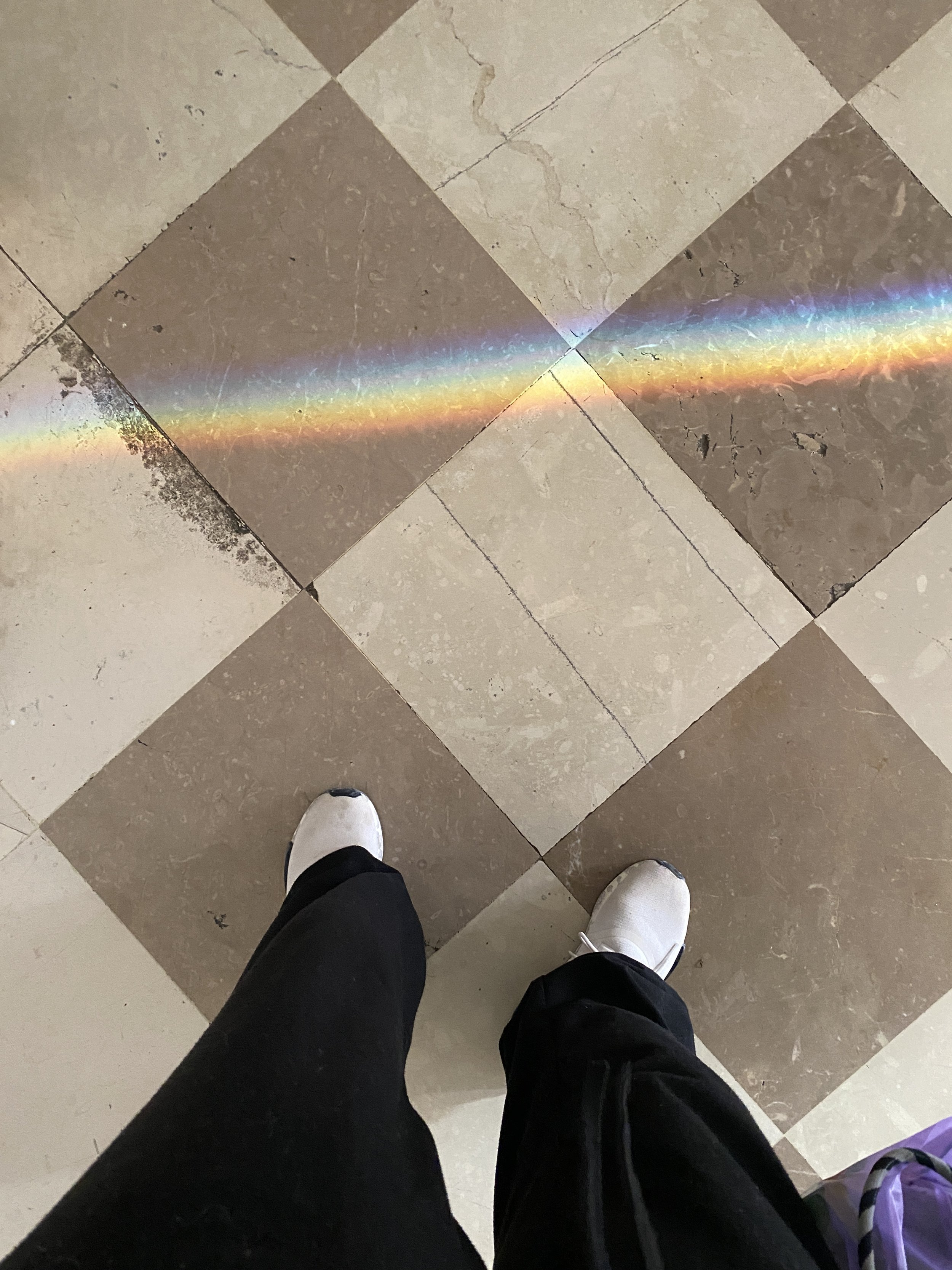 View of a tiled floor with a rainbow-colored light reflection across the center, with a person's legs and feet visible, wearing black pants and white shoes.