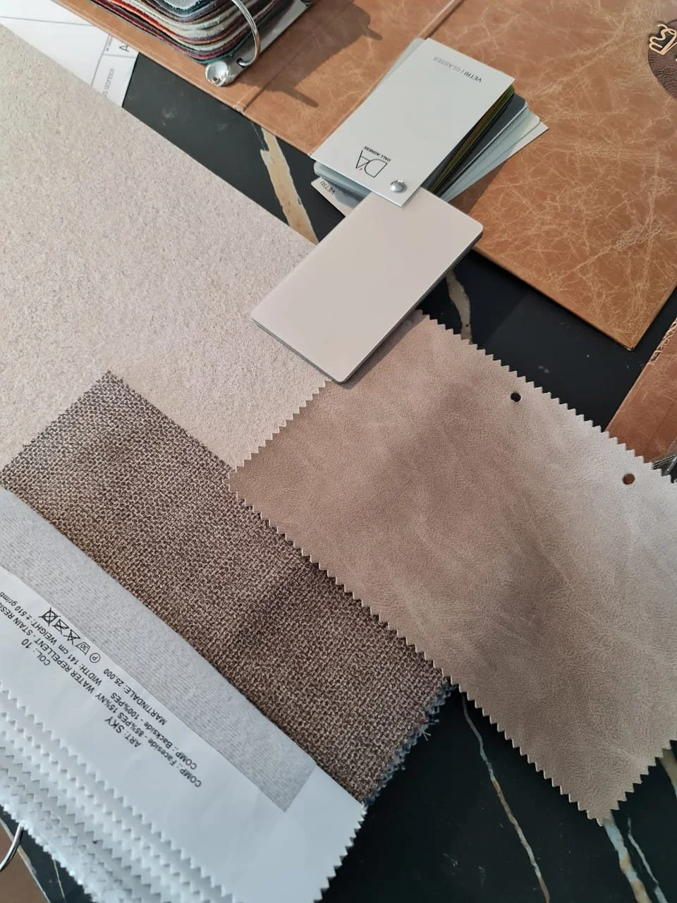 Samples of fabric swatches in beige, cream, and textured patterns placed on a black marble surface, alongside color and material sample cards and a small stack of color sample booklets.