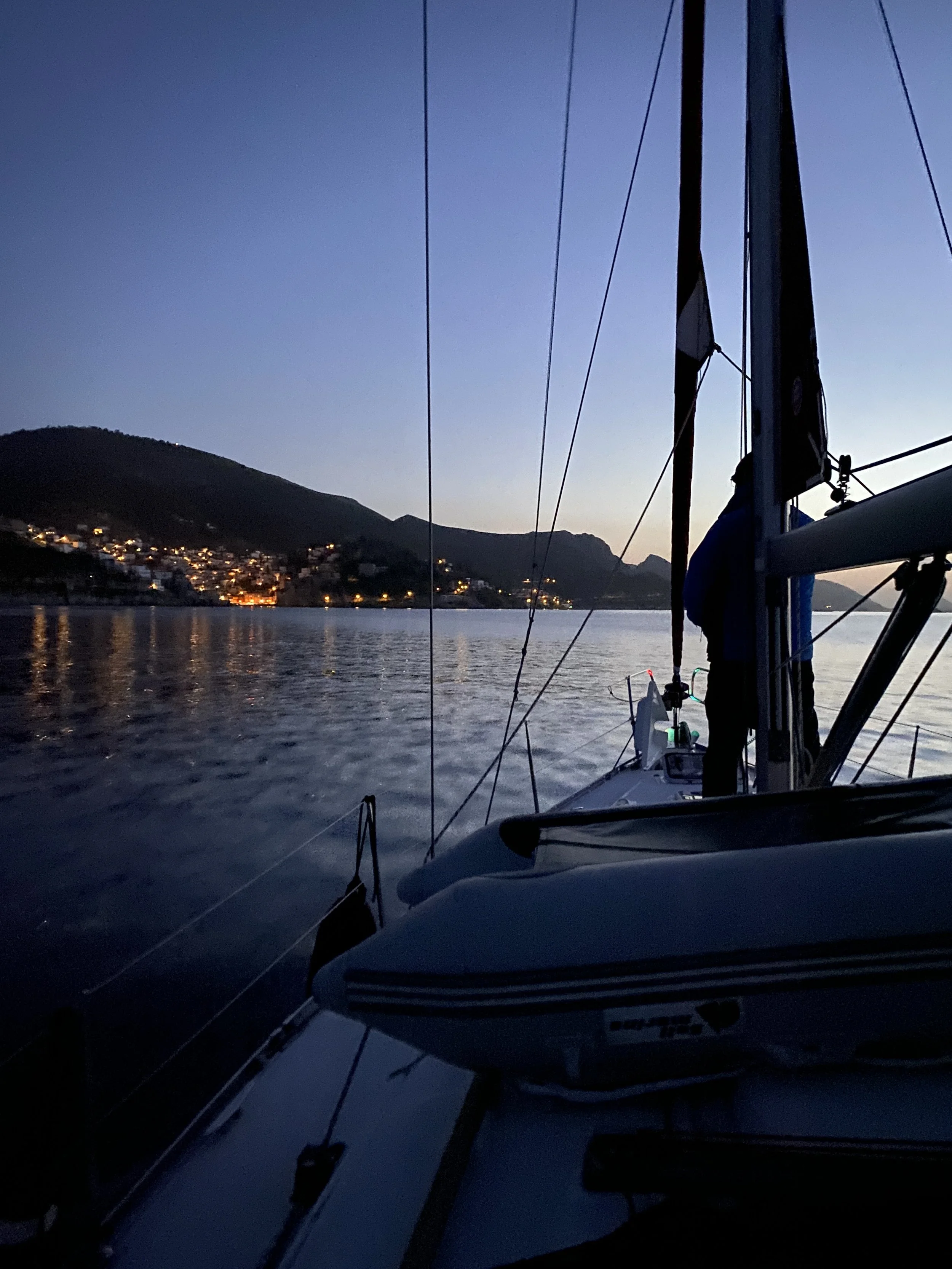 View of a sailboat on calm water during dusk with a mountain and a small town with illuminated houses in the background.