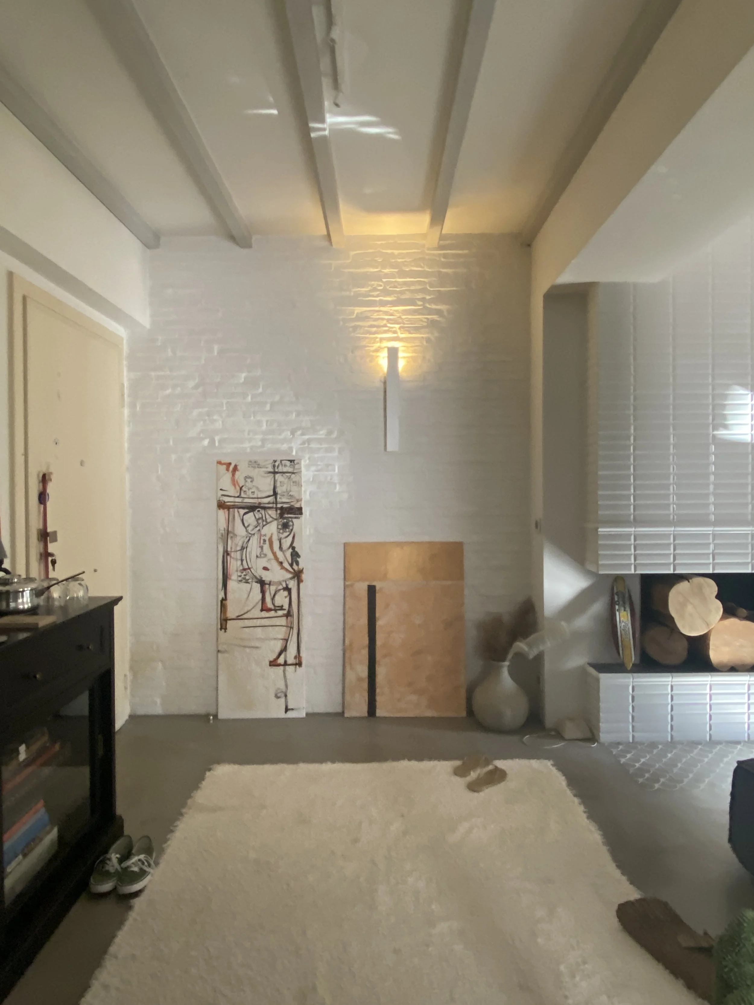 Living room with white brick walls, a white textured rug, and artwork leaning against the wall. A black window with blinds partially opened is on the right, with logs placed near the fireplace. A small table with books and shoes are on the left, and 