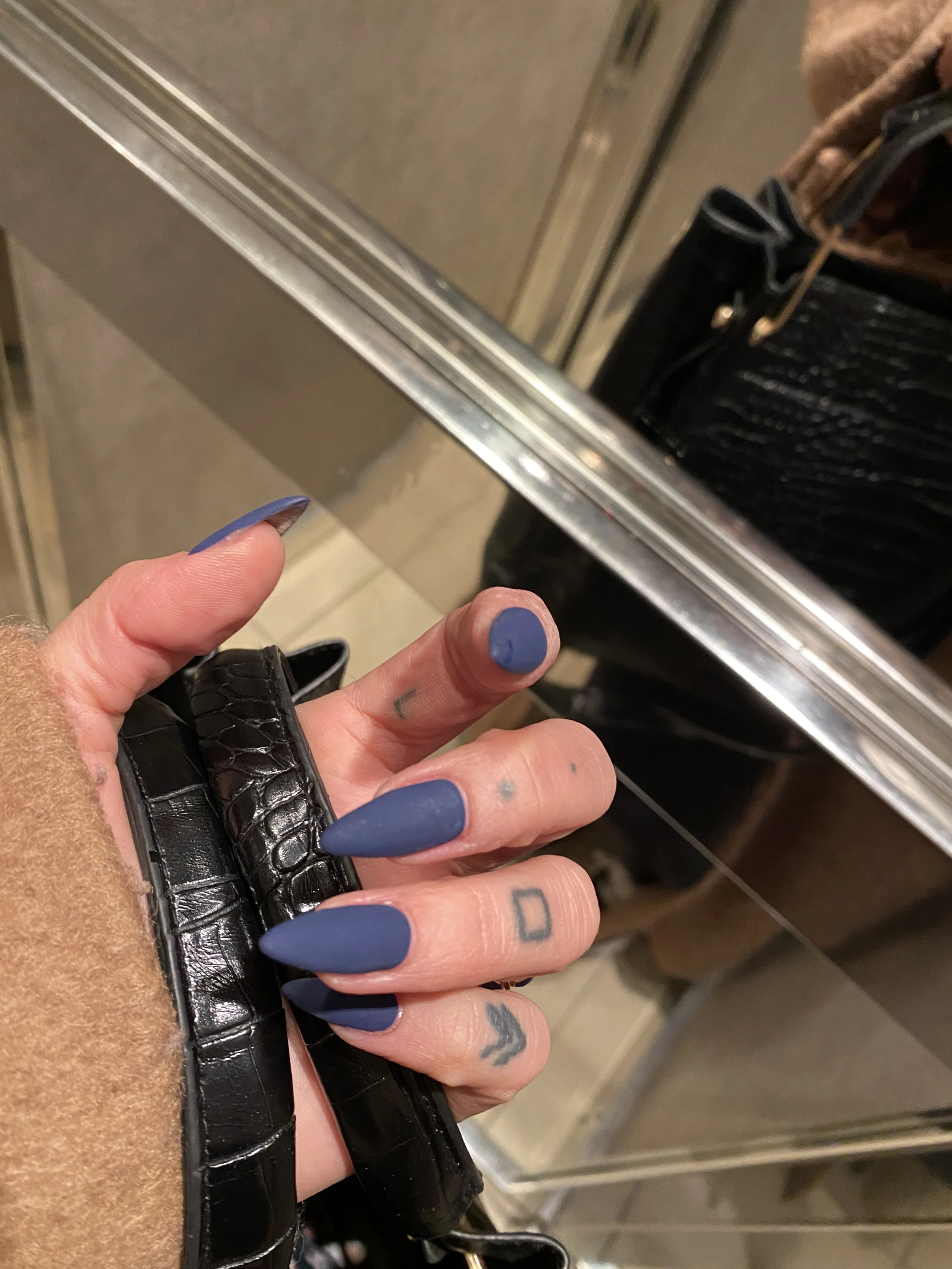 A person's hand with multiple tattoos holding a black purse with a crocodile skin pattern. The hand has manicured nails painted in matte blue. The background includes a metallic surface and part of a metal shopping cart or luggage.