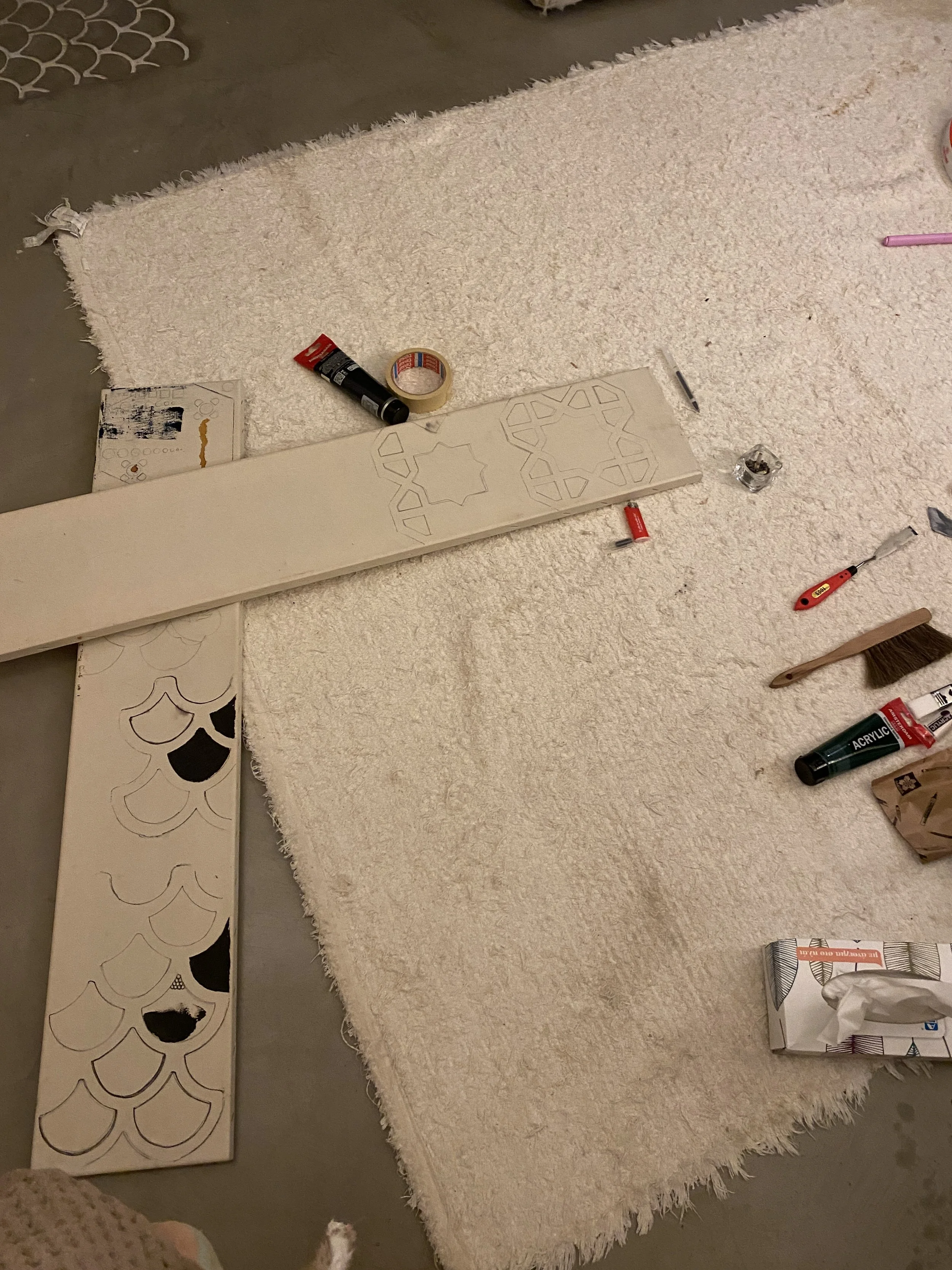 Craft workspace on a beige rug with stencil patterns, paint, a brush, electrical tape, a screwdriver, and other tools scattered around.