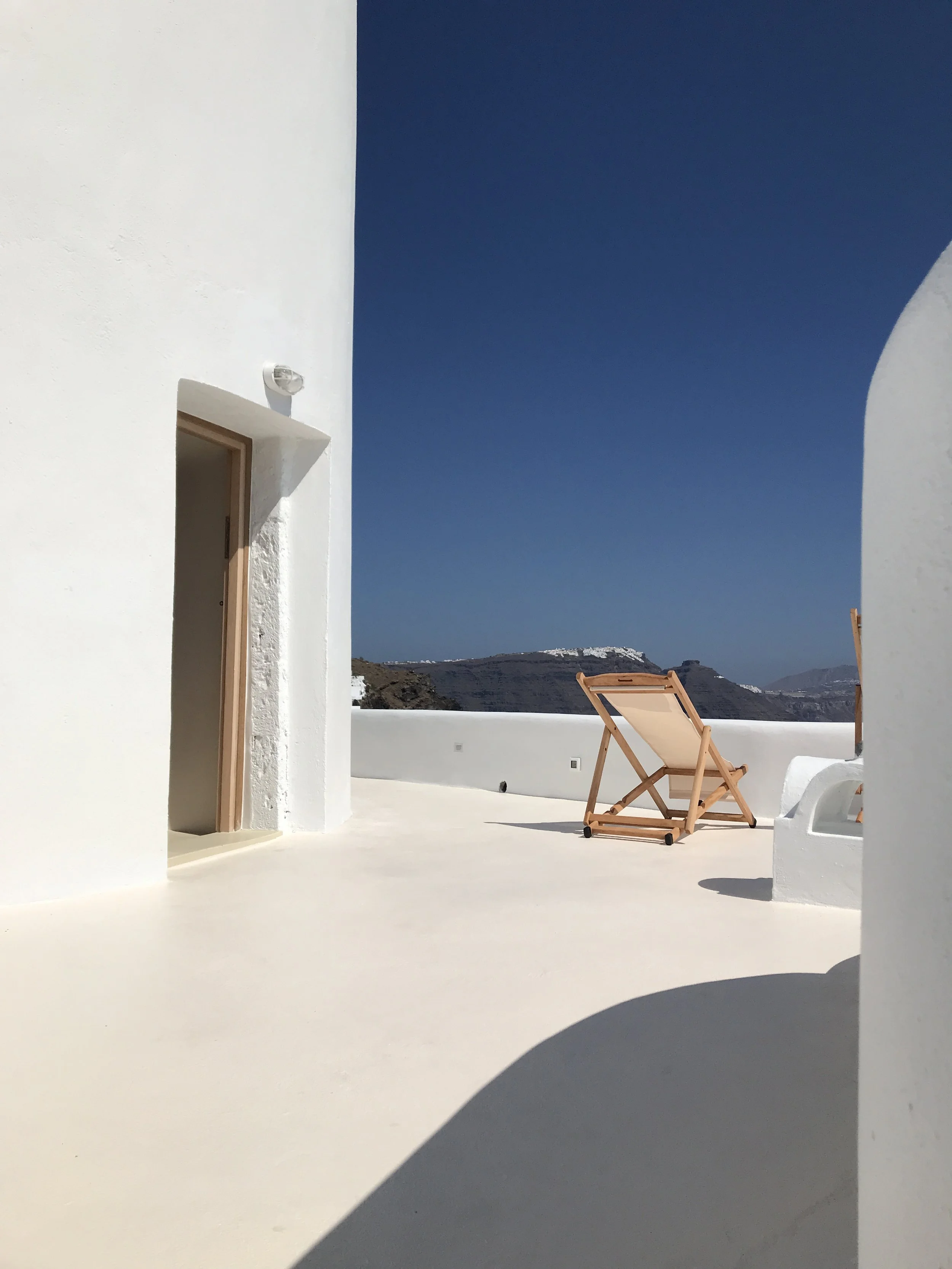 White building with a wooden door, a beige deck chair on a white terrace, blue sky, and distant mountainous landscape.