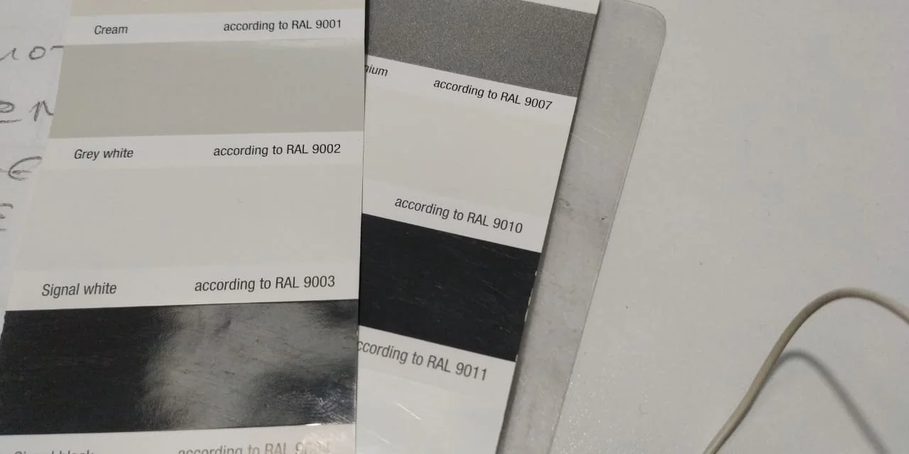 Two color sample cards showing different shades of gray and white with their RAL codes, placed on a light-colored surface, along with a handwritten note partially visible on the left.