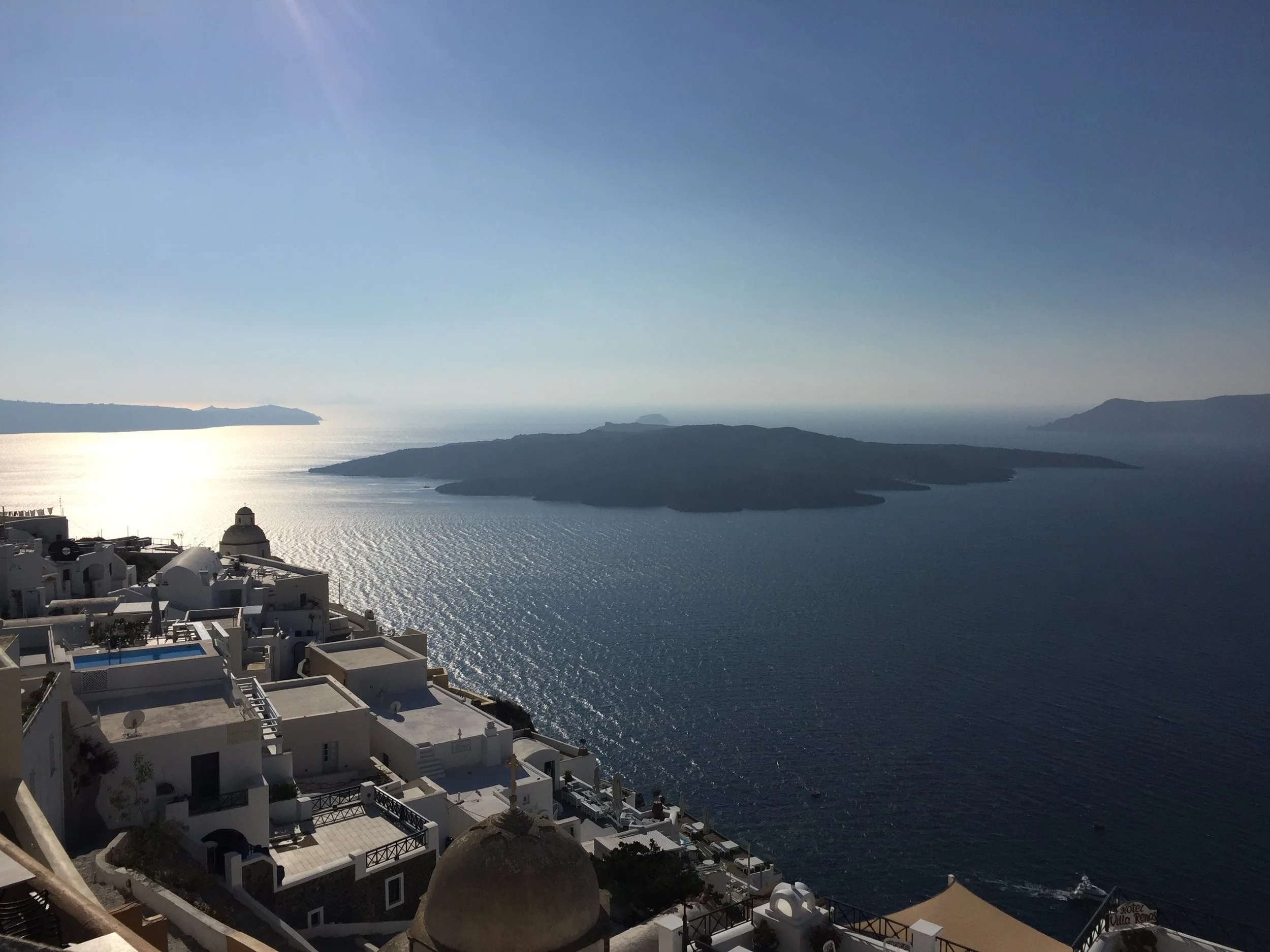 White buildings with flat roofs and domed structures on a hillside overlooking a large body of water, with islands and distant landforms under a clear sky.