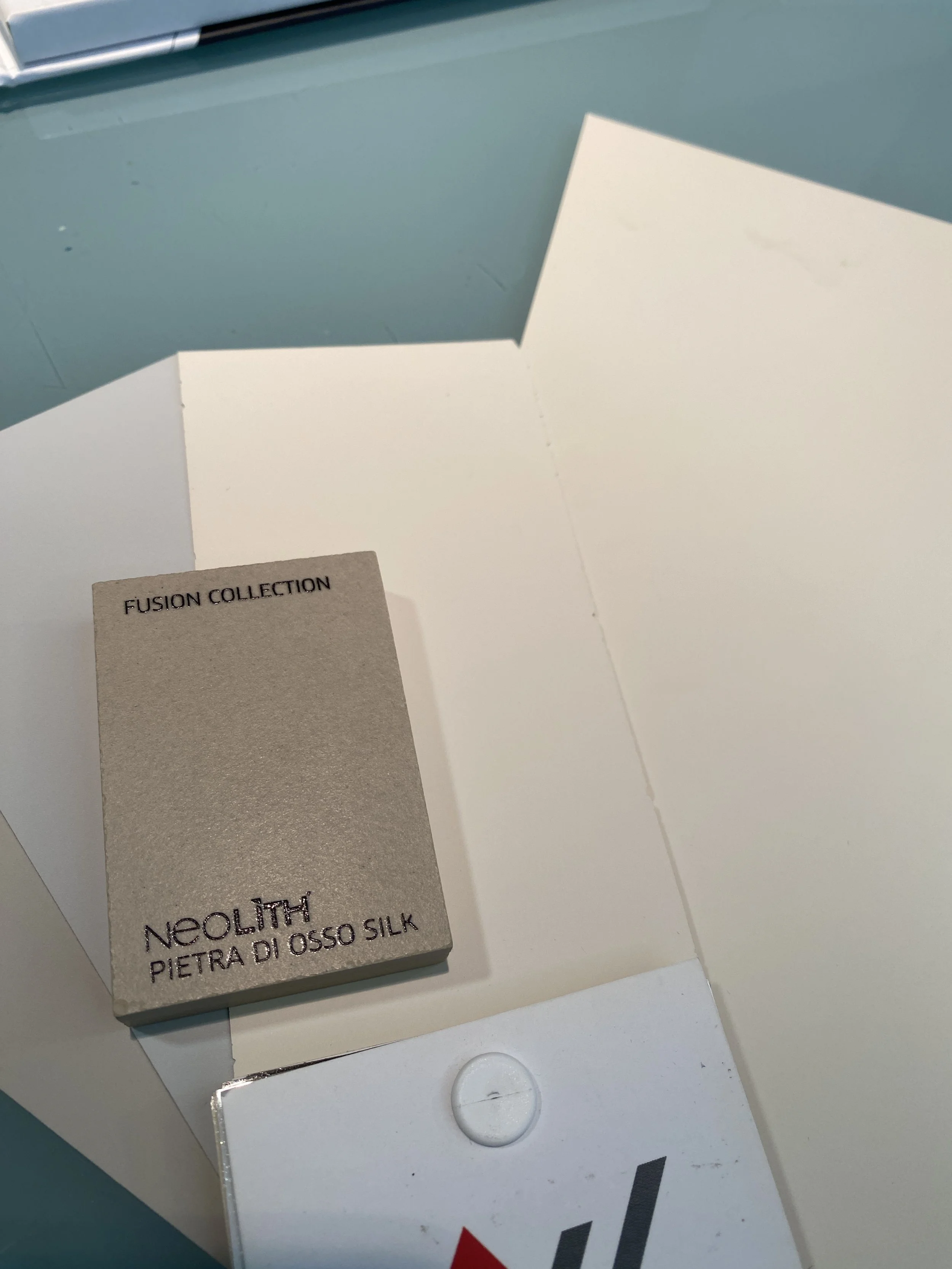 A stack of Neolith Pietra di Osso Silk sample cards on white paper. The cards have a grey textured surface and black text. The background shows a blue surface and part of a metallic object.