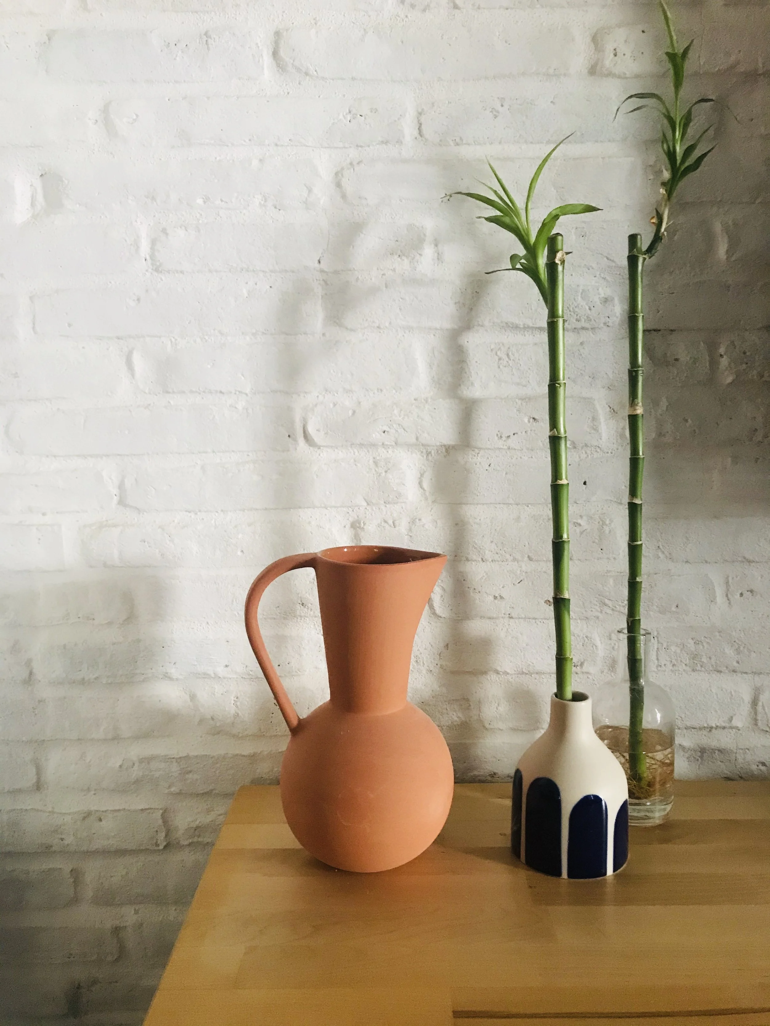 A pink pitcher with a handle on a wooden table. Behind it, two bamboo stalks in white and clear glass vases. To the right, a white vase with black designs. The background features a white brick wall.