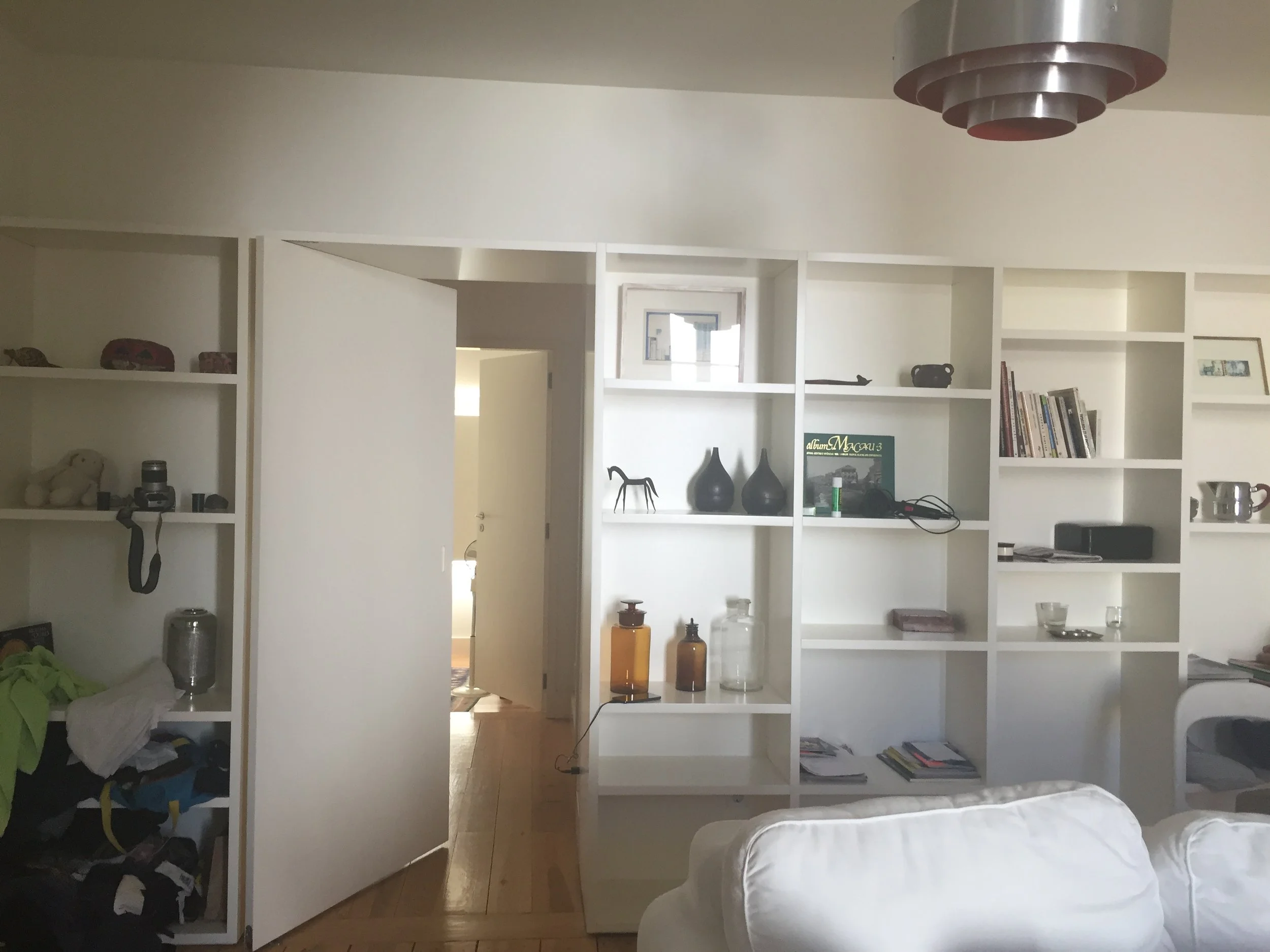 Living room with white built-in bookshelf and door open to another room, with various decorative items, books, and clutter visible.