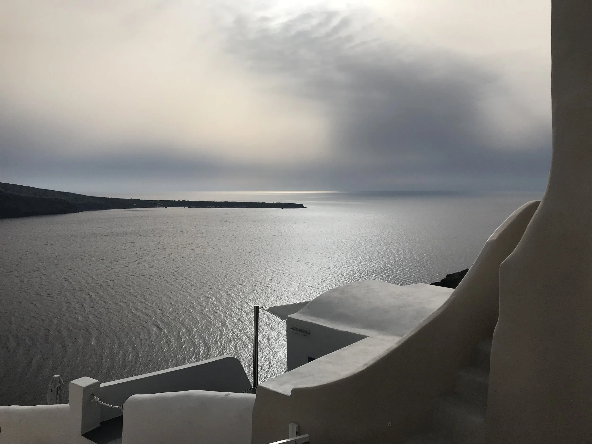 View of a coastal landscape with the sea, a rocky shoreline, and cloudy sky during daytime, taken from a white architectural structure, possibly a balcony or terrace.