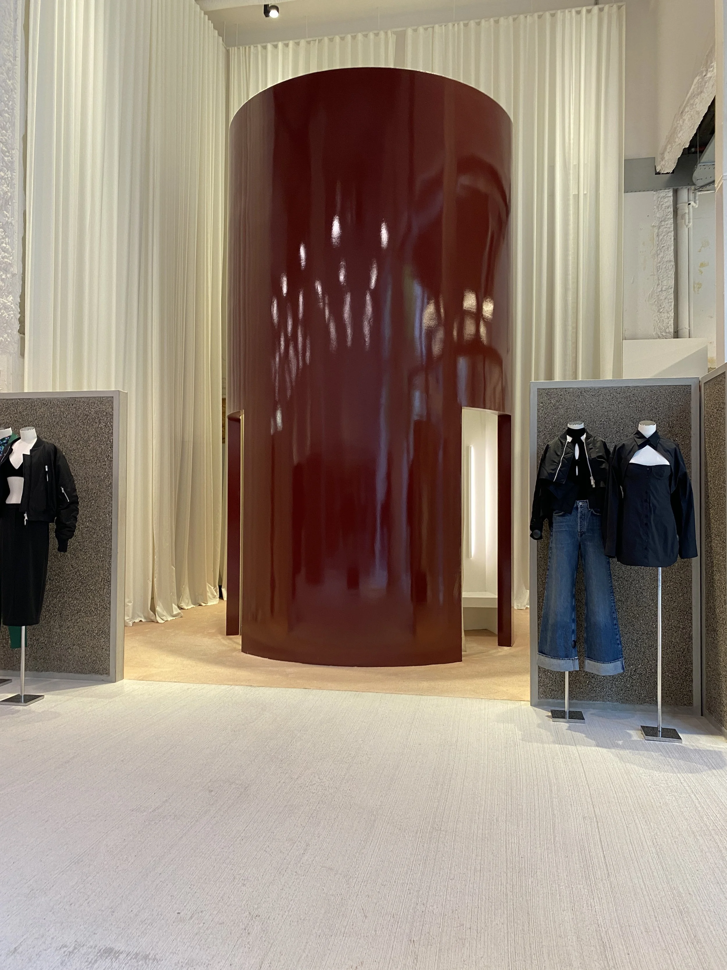 Fashion display in a store with mannequins dressed in black and white clothing, set against grey partitions and a large, curved, shiny red wall in the background.