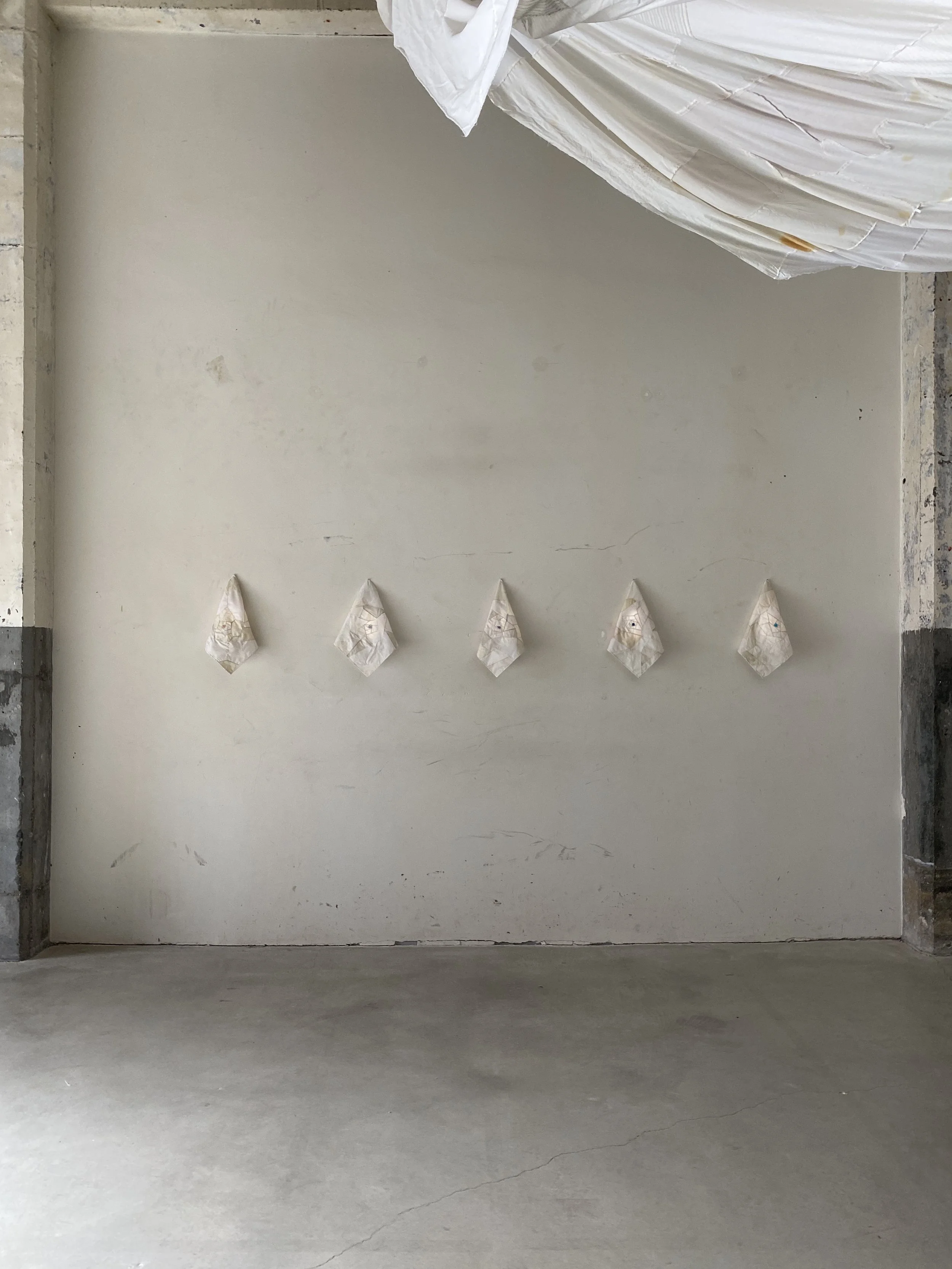 A plain white wall with five paper bags or pouches attached across the middle, evenly spaced. In the top right corner, part of a white fabric or sheet is draped, partially visible.