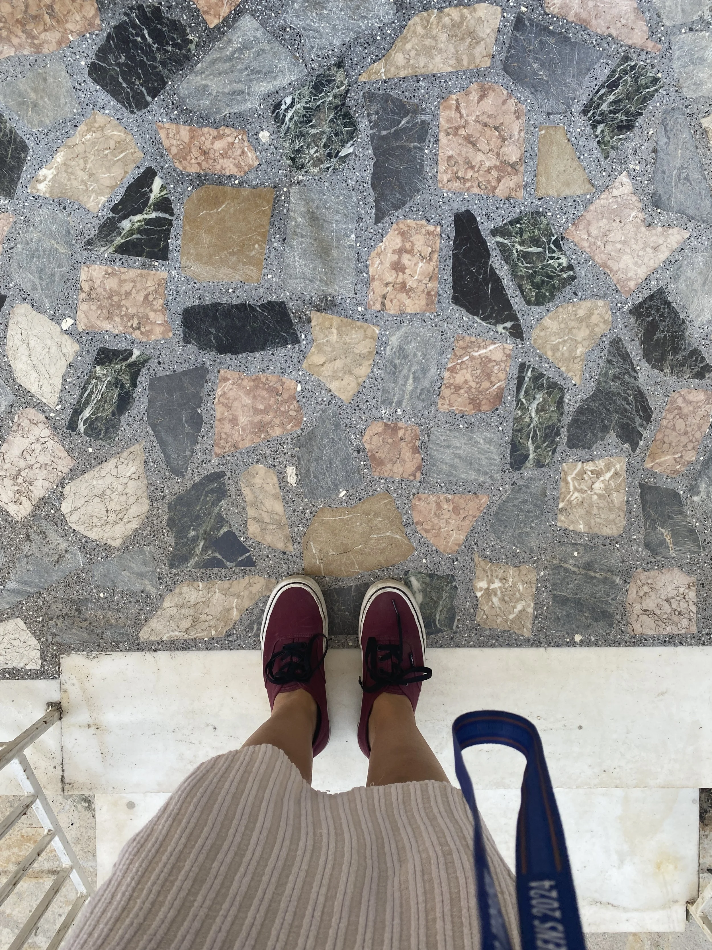 A person's legs wearing maroon sneakers with black laces and a beige striped dress, standing on a multicolored stone floor near a white marble step and holding a blue lanyard.