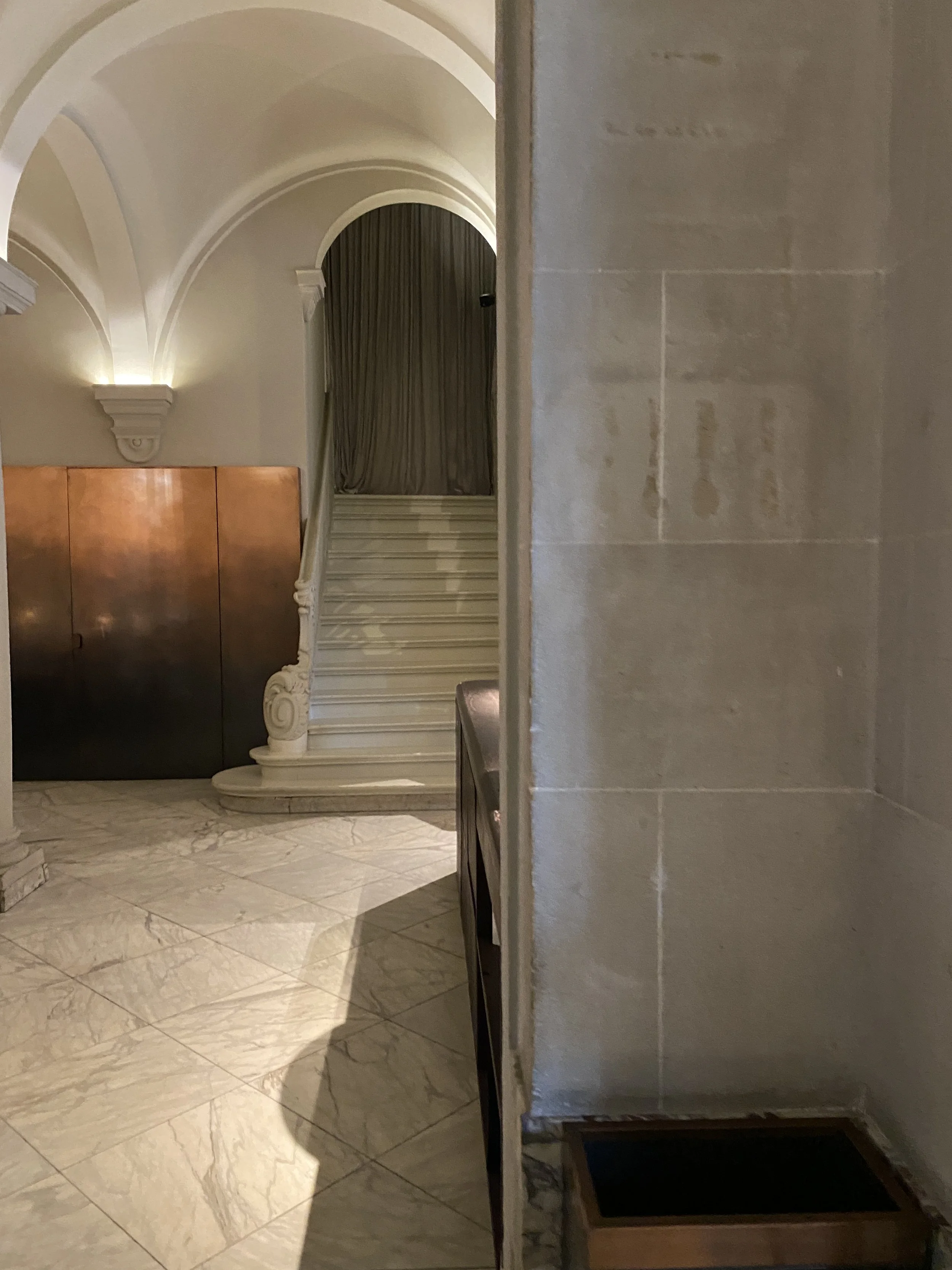Entrance hall of a building featuring marble tiled flooring, a staircase with ornate details, a curtain at the top of the stairs, and a flat wooden door.