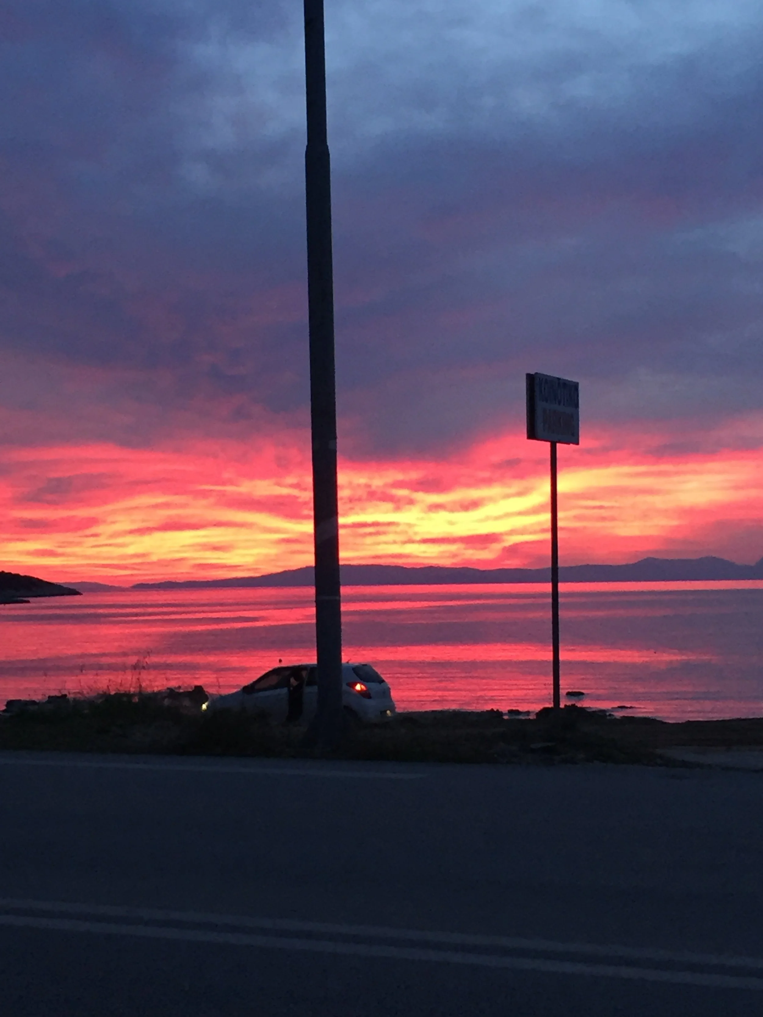 Colorful sunset over a body of water with a car parked near the shoreline and street signs visible in the foreground.