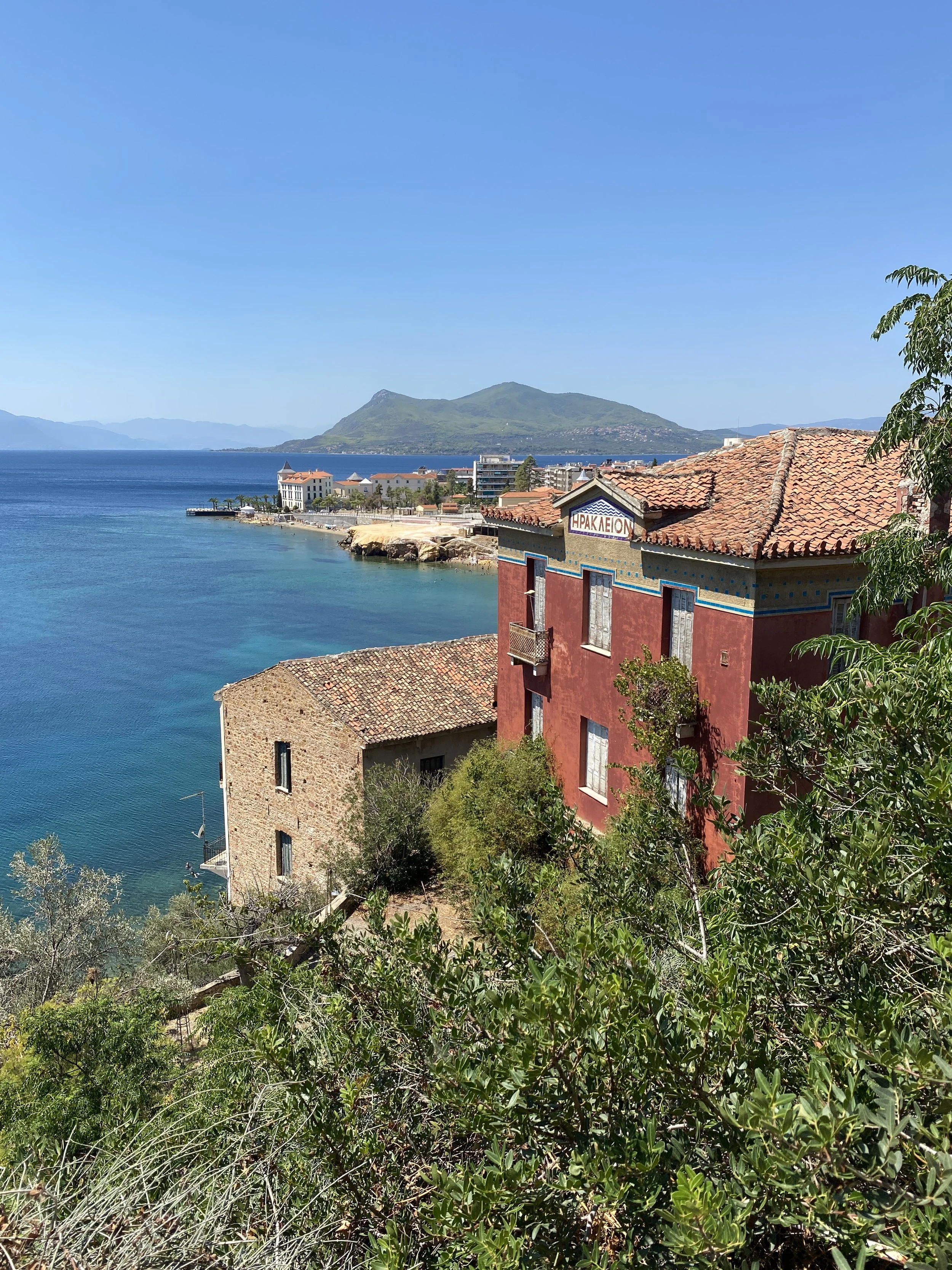 View of a coastal town with red and beige buildings, a calm blue sea, and green mountains in the background. Clear sky.