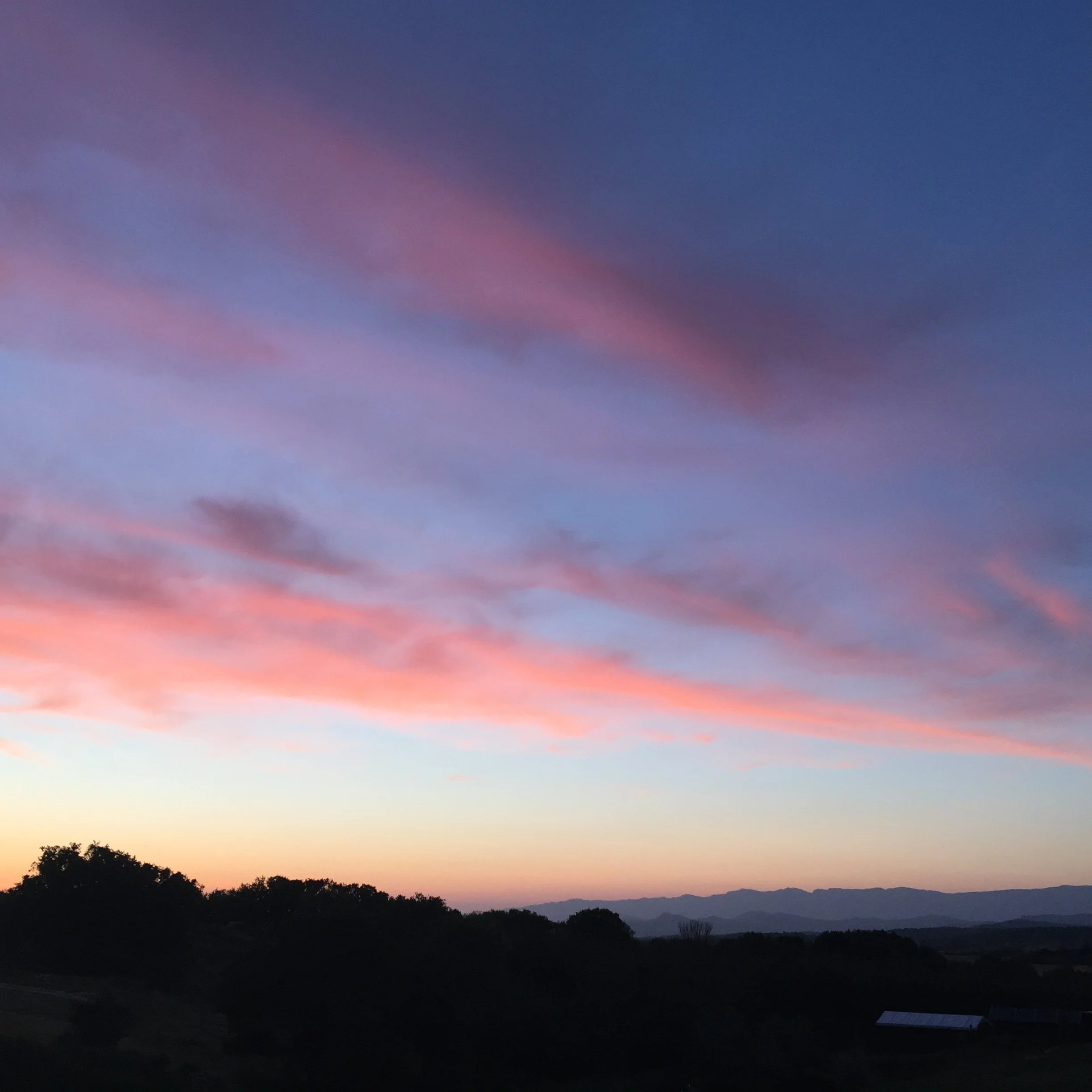 A landscape of trees and mountains at sunset with a colorful sky in shades of orange, pink, purple, and blue.