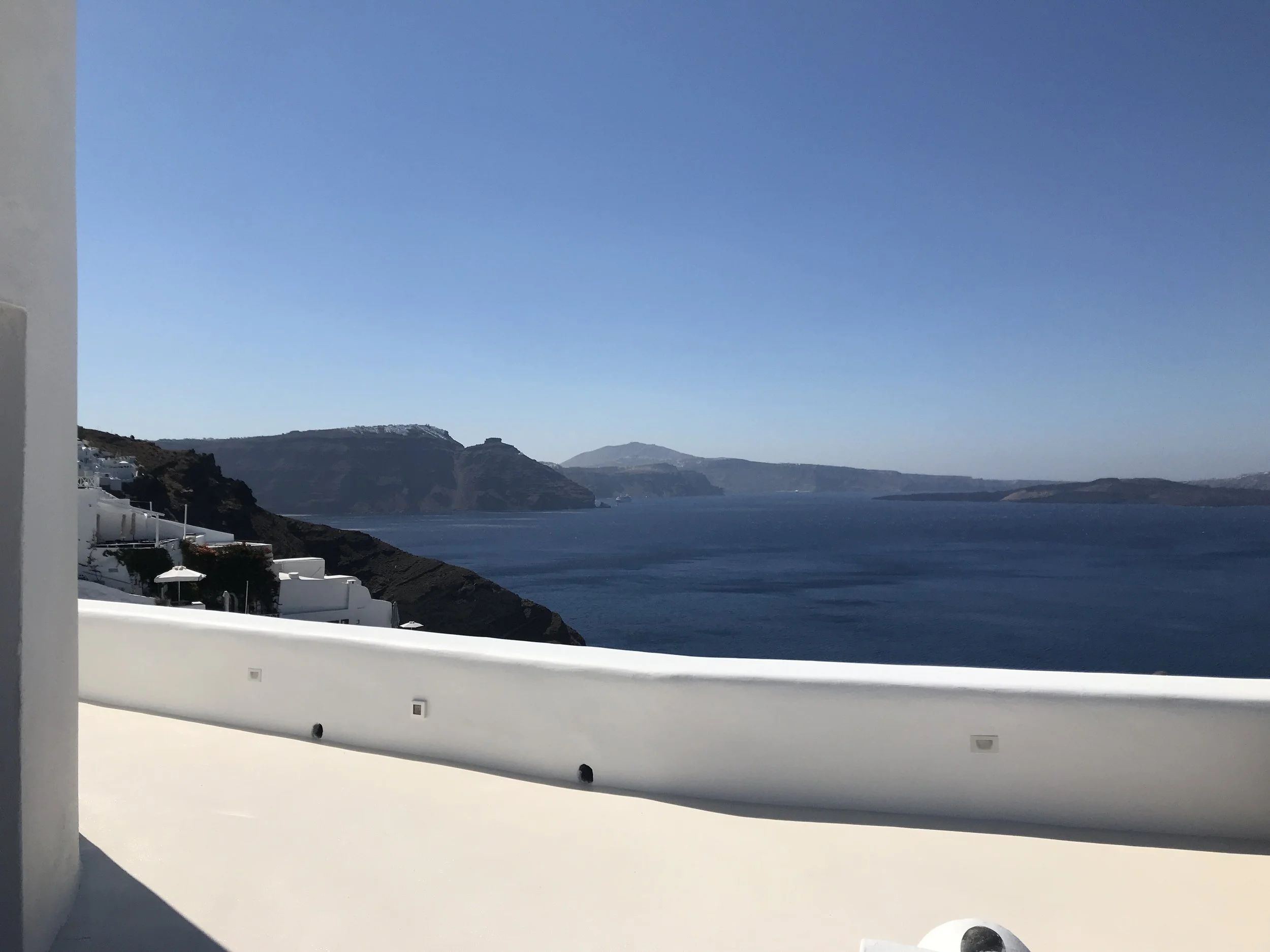 A picturesque view of a blue sea and distant hills under a clear sky, partly visible from a coastal terrace with white walls.
