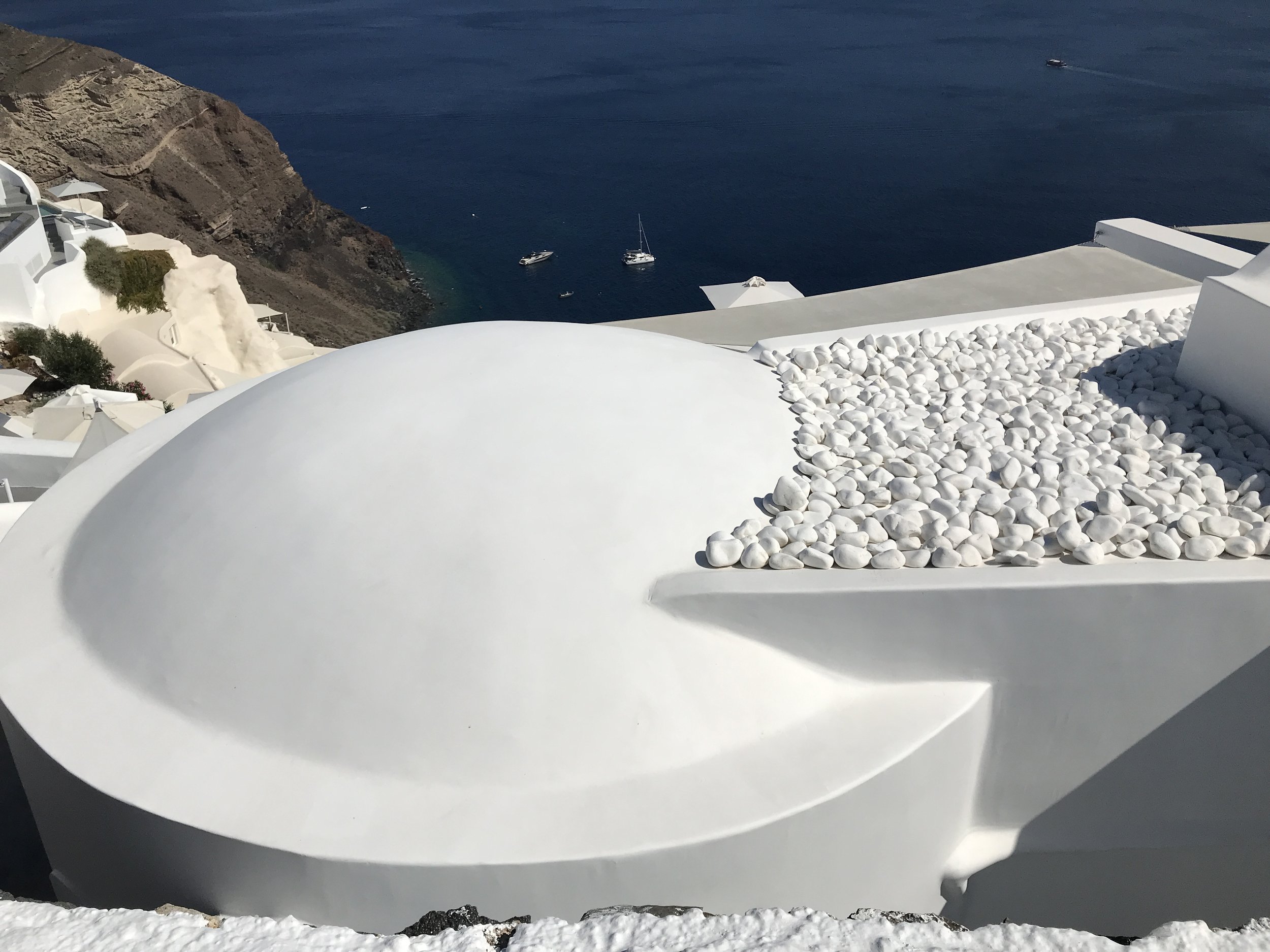 White building with smooth curved roof and a section filled with white stones, overlooking a blue sea with boats, on a steep hillside
