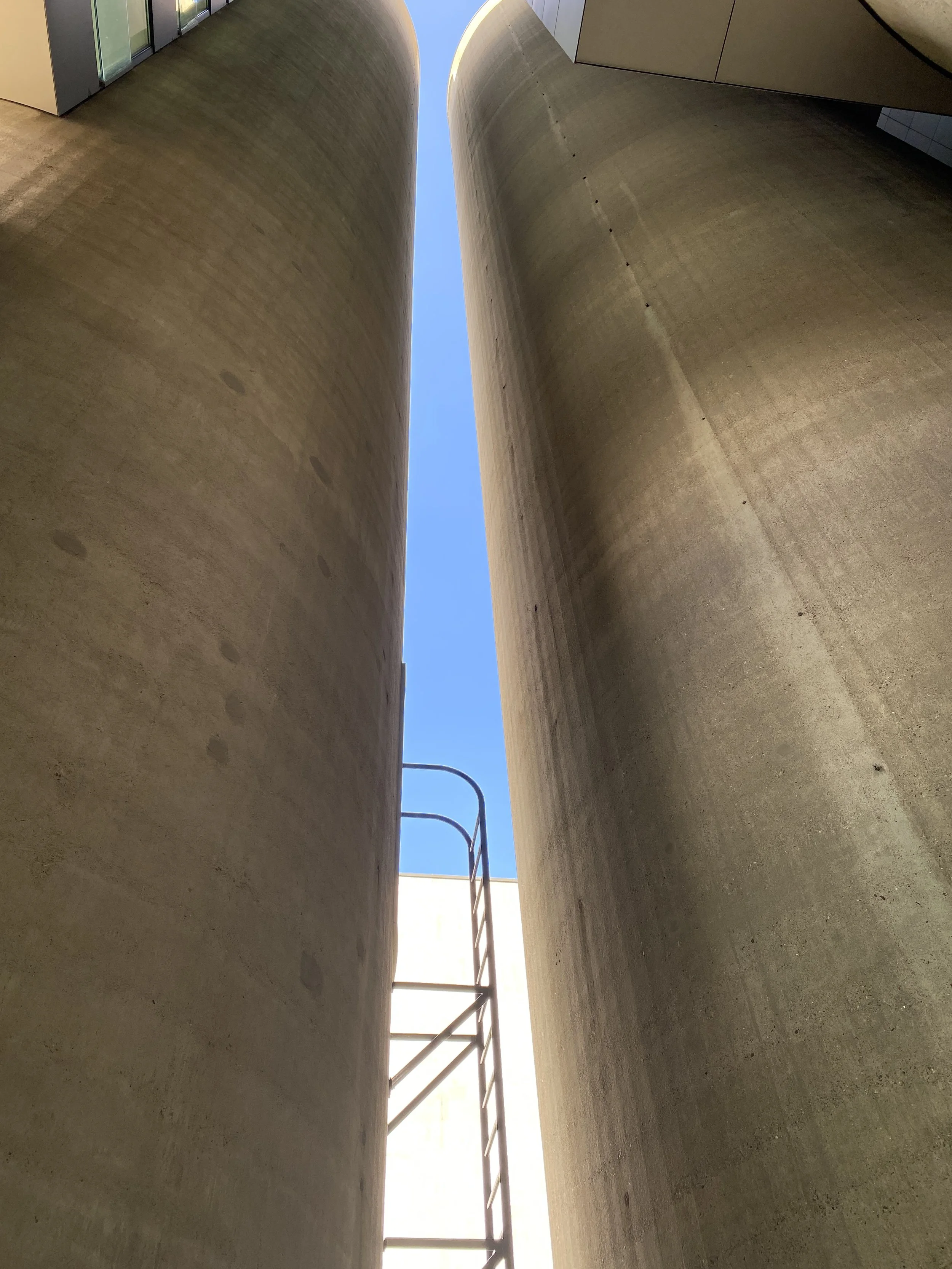 Looking up between two large concrete silos with a small ladder and blue sky visible at the top.