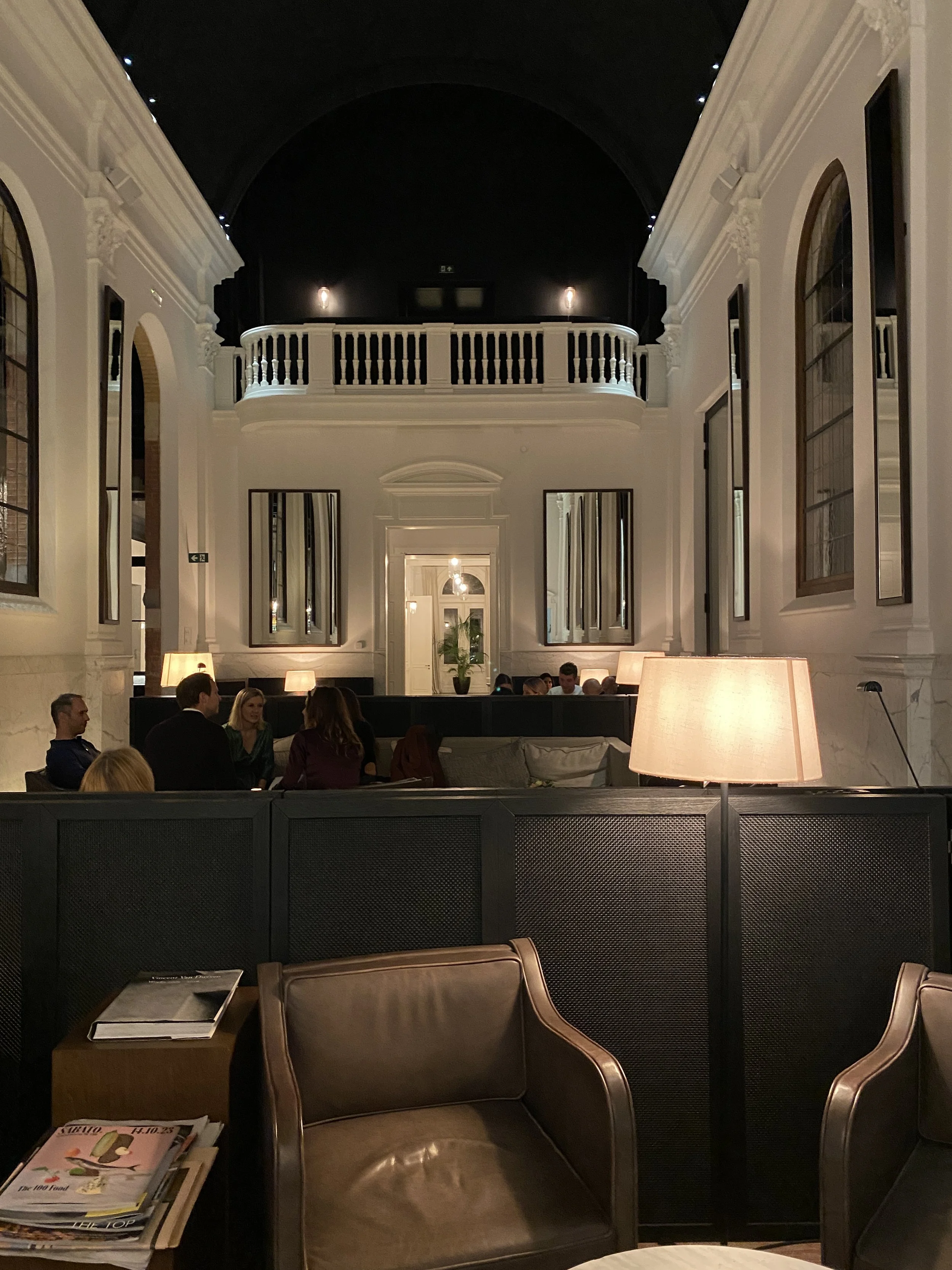 Interior of a sophisticated lounge or restaurant with high ceilings, arched windows, and warm lighting. People are seated at tables and sofas, engaged in conversation. Classic decor with mirrors and lamps.
