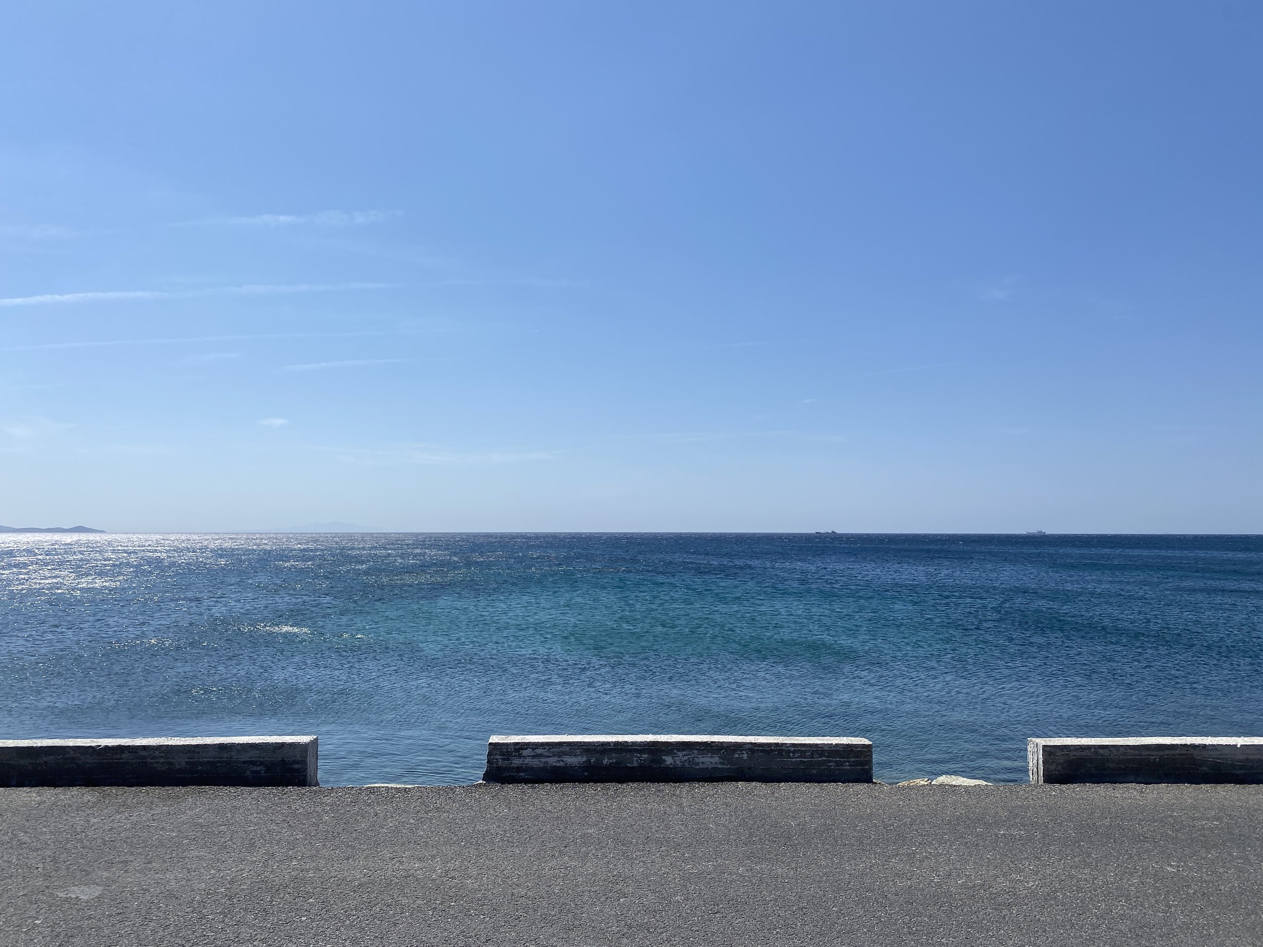 View of the ocean with a clear blue sky, reflecting sunlight on the water, and a concrete barrier in the foreground.