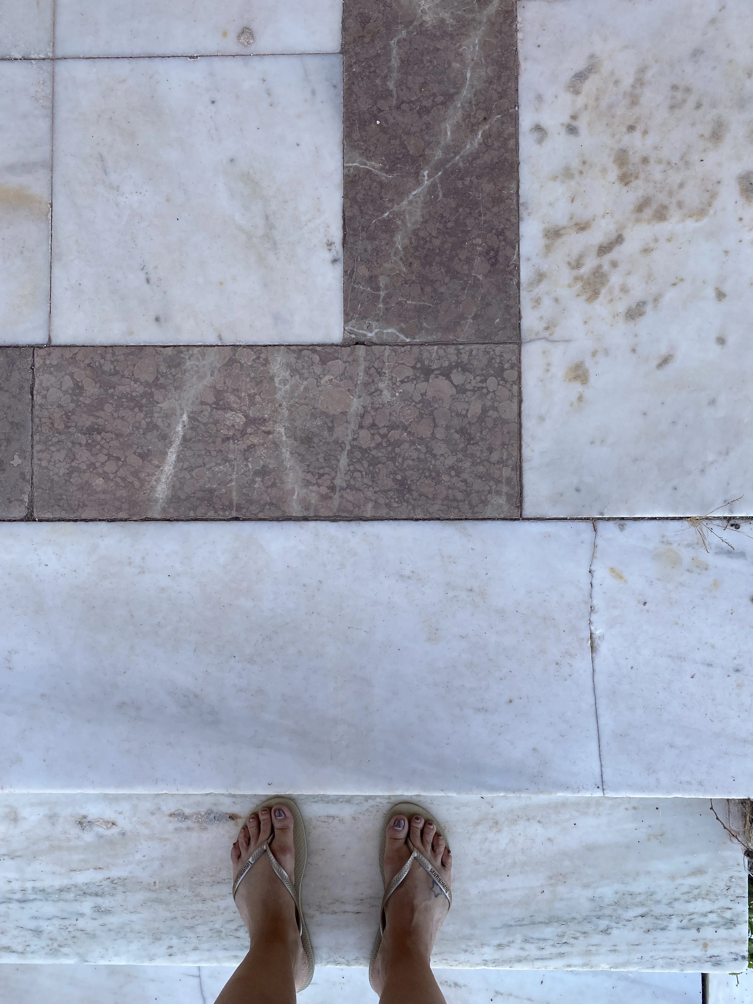 Photo taken from above of a person's feet in sandals on a tiled floor with a pattern of white and brown tiles.