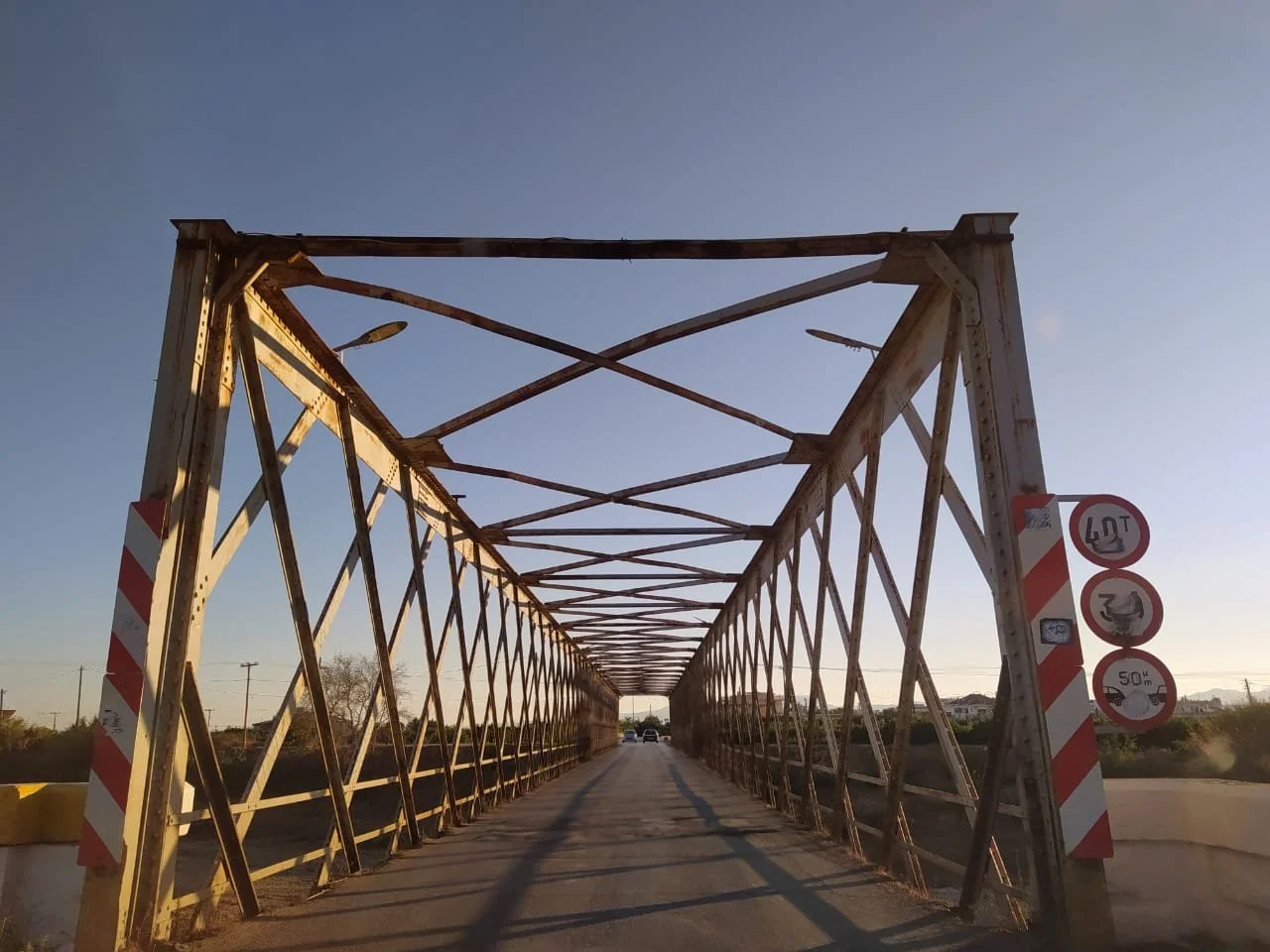 An old metal truss bridge with rust and peeling paint, spanning a roadway with cars. Signage indicates height and weight limits, and no overtaking. The sky is clear with a soft sunset glow.