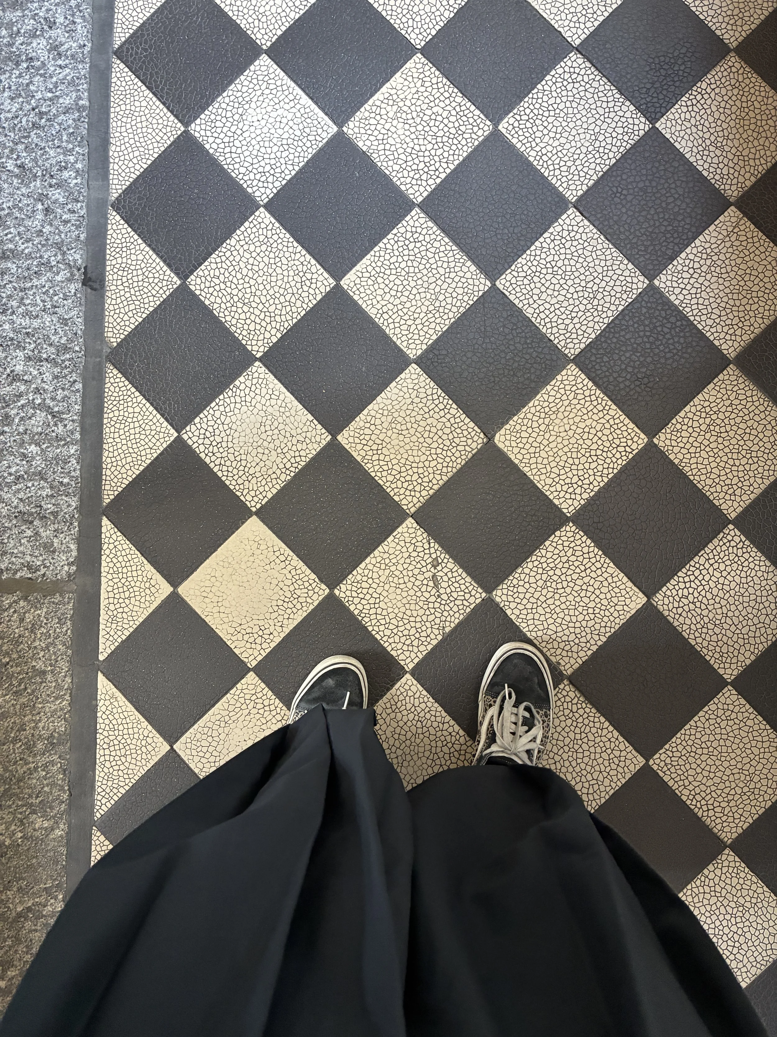 Photo of a person's black shoes and black pants standing on black and beige checkered tile floor.