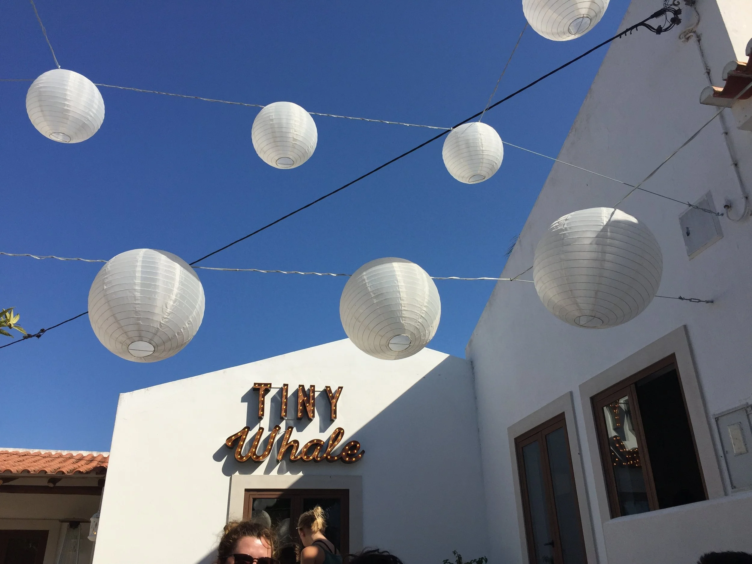 White paper lanterns hanging against a bright blue sky above a white building with a sign that says 'TINY Whale' and people in the foreground.