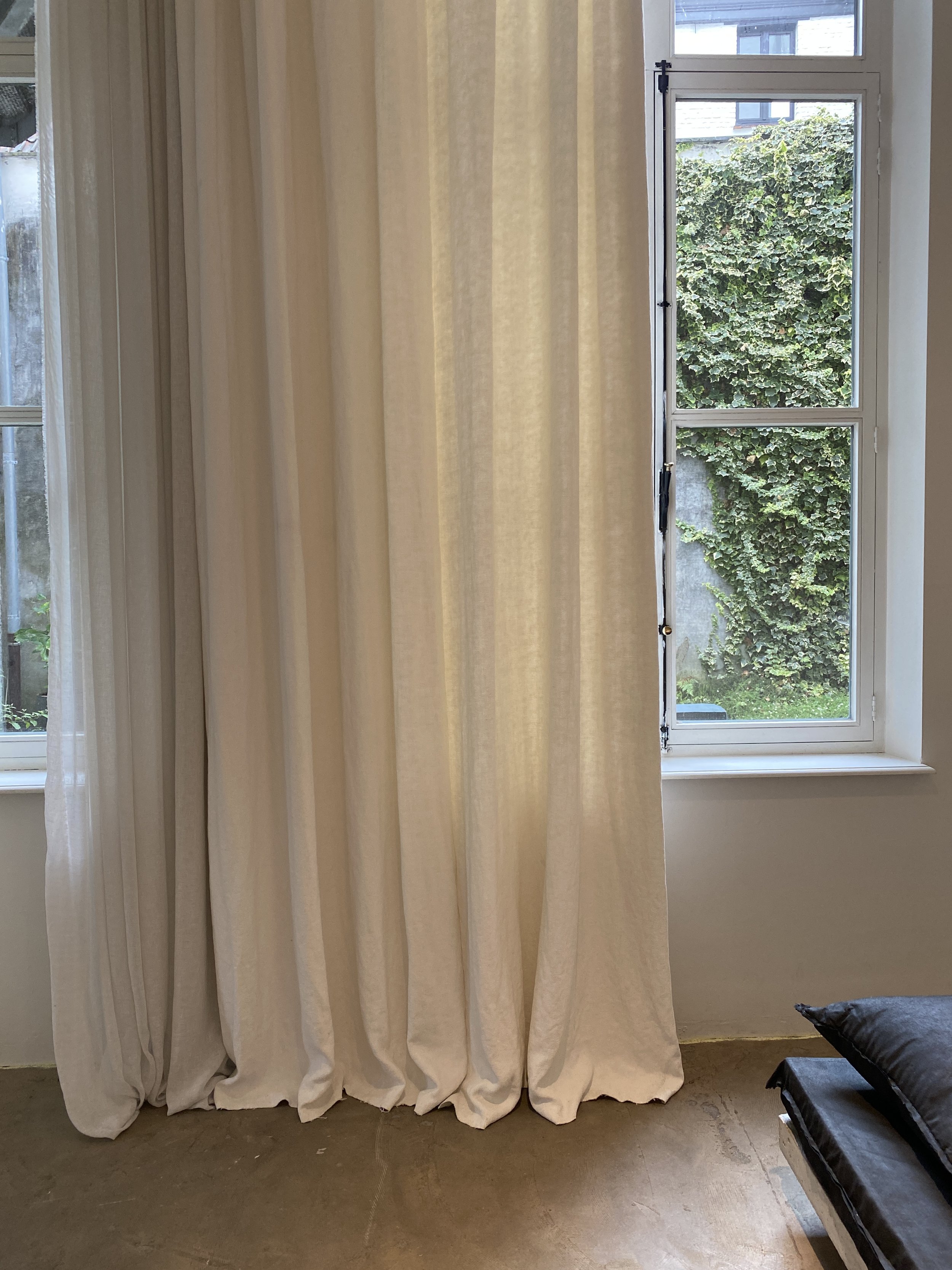 A window with white frame and glass panes, showing green foliage outside. Cream-colored curtains partially cover the window, and a black sofa with a pillow is visible in the room.