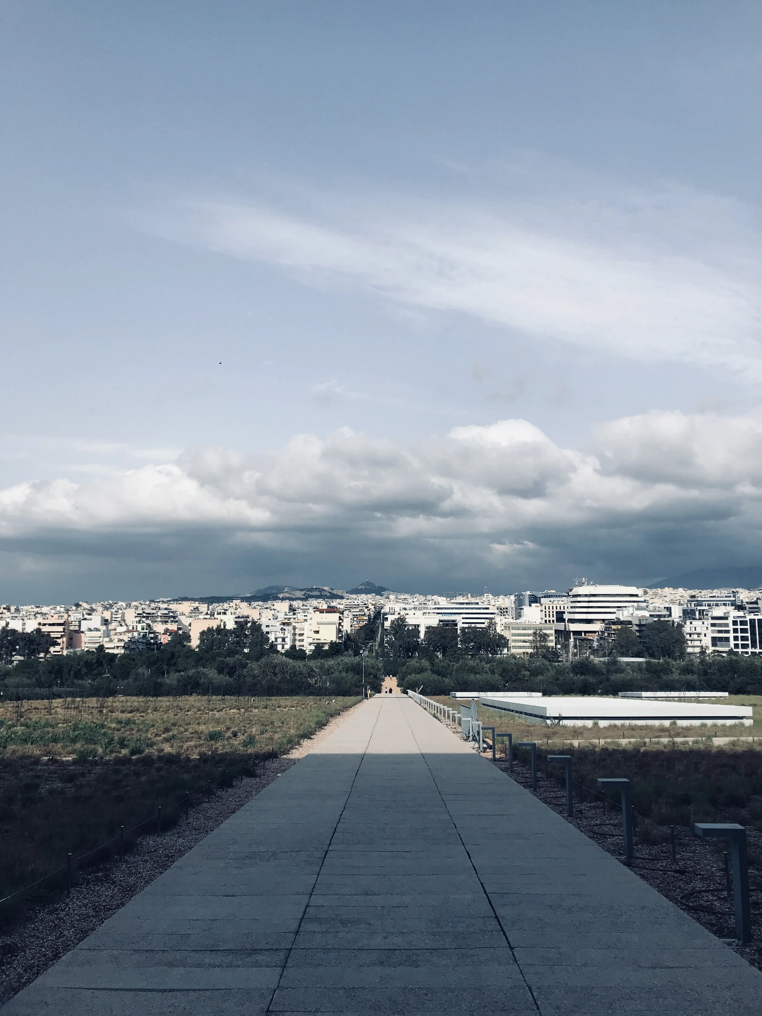 A wide view of a cityscape under a partly cloudy sky with a concrete sidewalk leading into the city, surrounded by greenery and modern buildings in the distance.