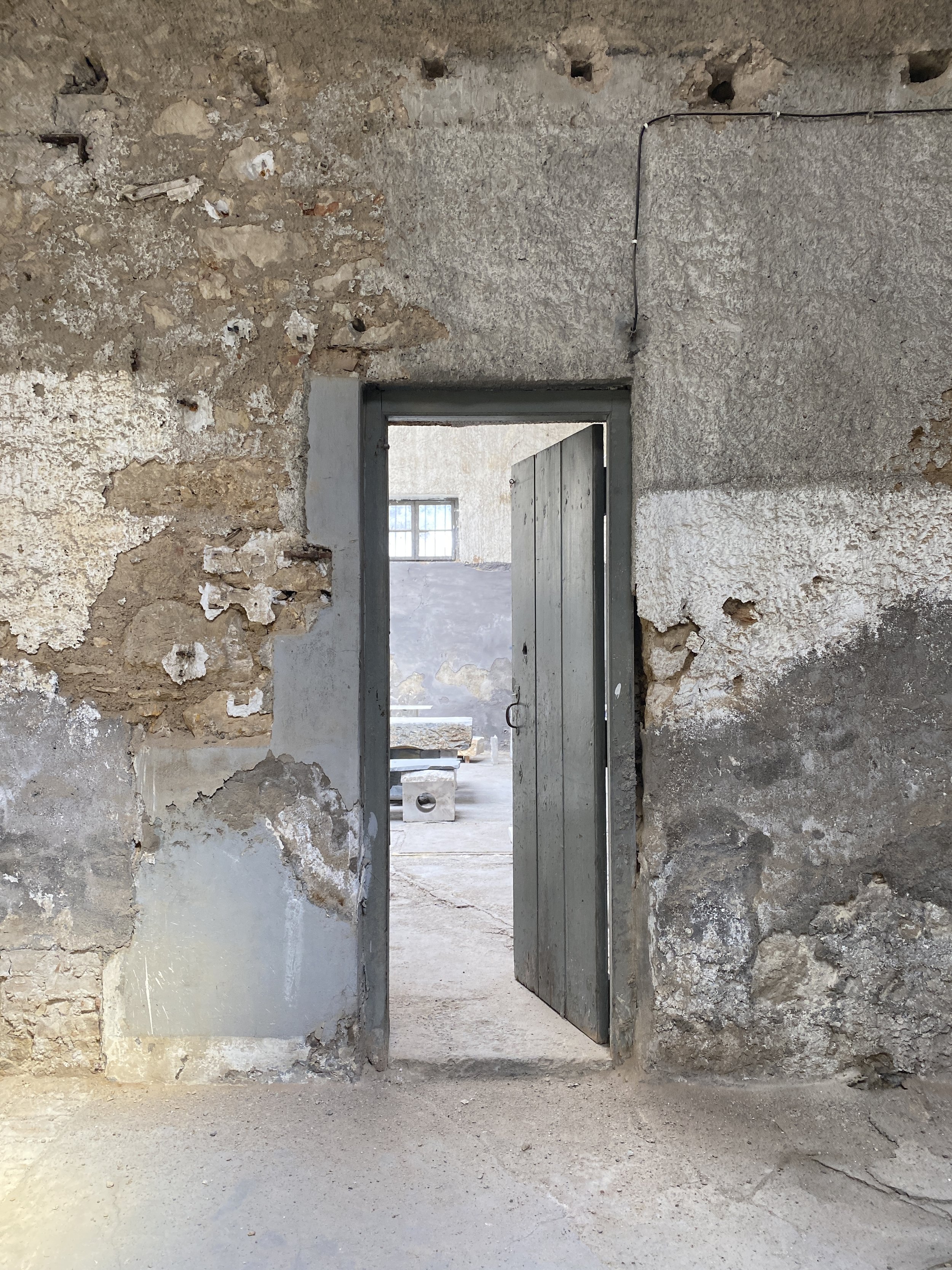 A partially open metal door in an old, worn stone and concrete wall, leading to an unfinished room with a window and construction materials inside.