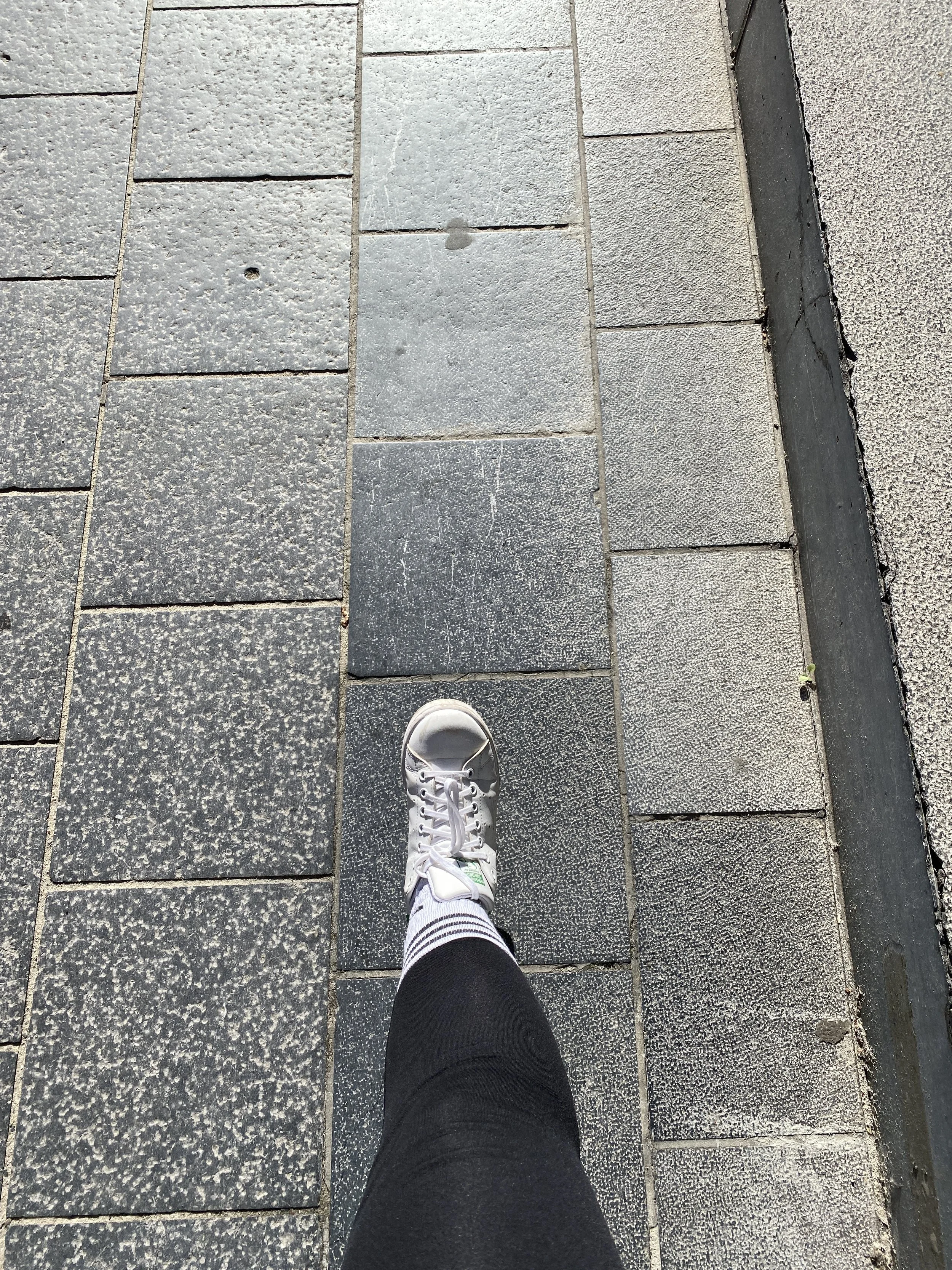 Top-down view of a person's foot wearing a white sneaker and striped sock walking on a sidewalk with rectangular gray paving stones.