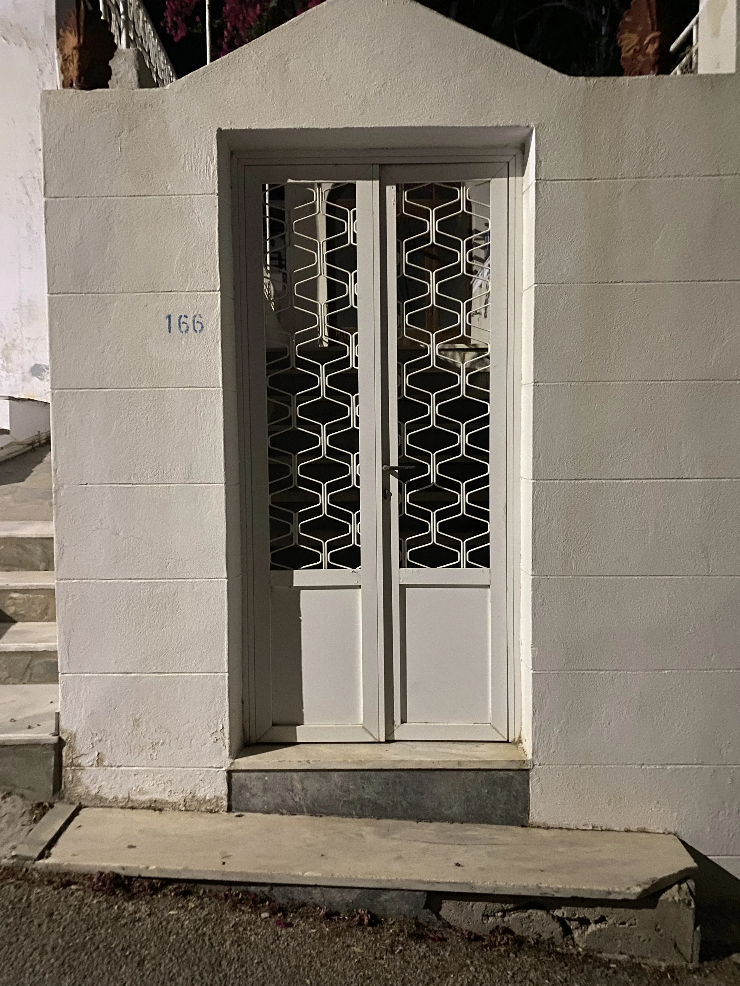 White front door with decorative metal grille, set in a white stucco wall, with the number 166 painted on the left side of the wall.