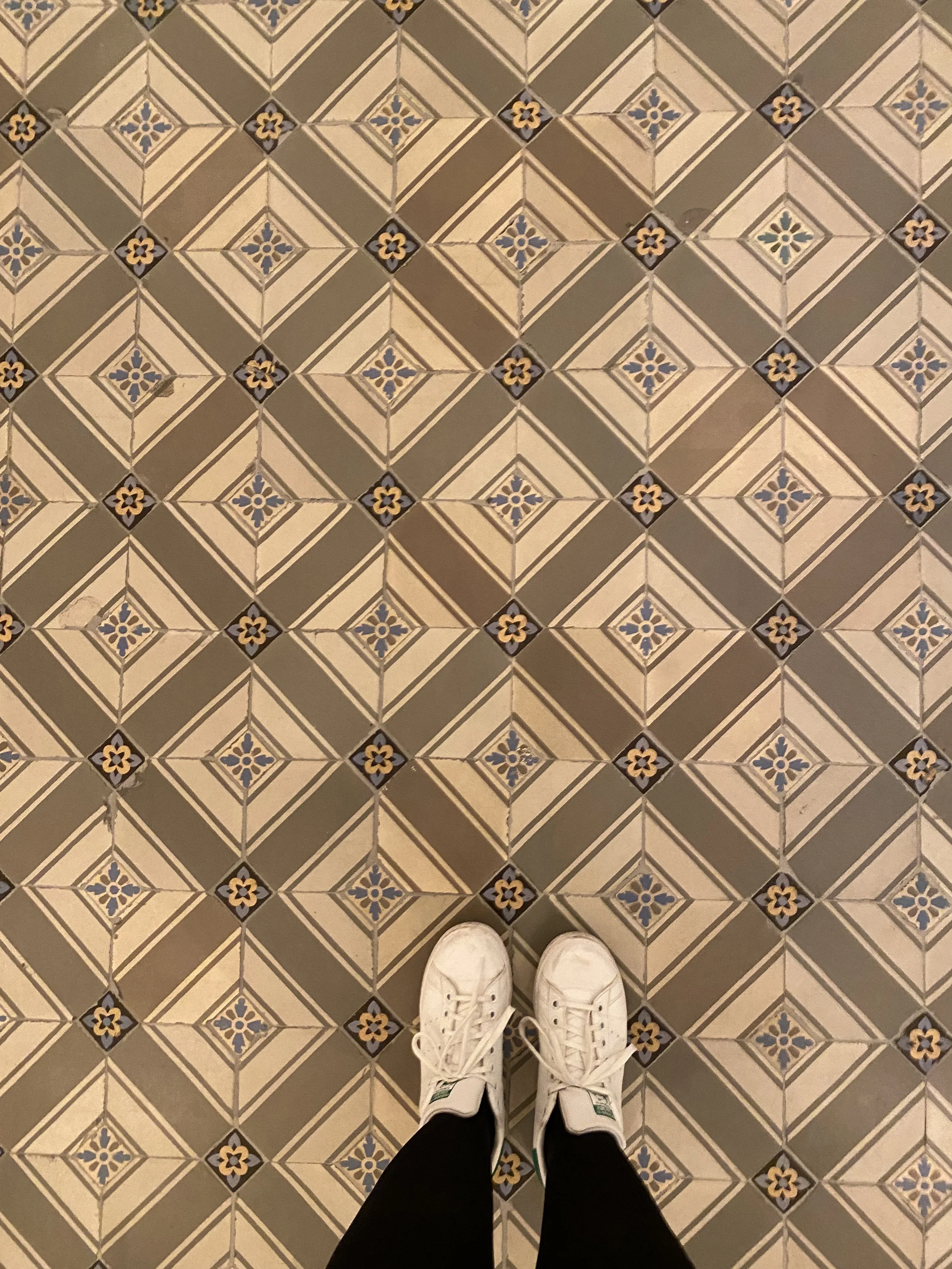 A person's white sneakers on a patterned tile floor with black pants.