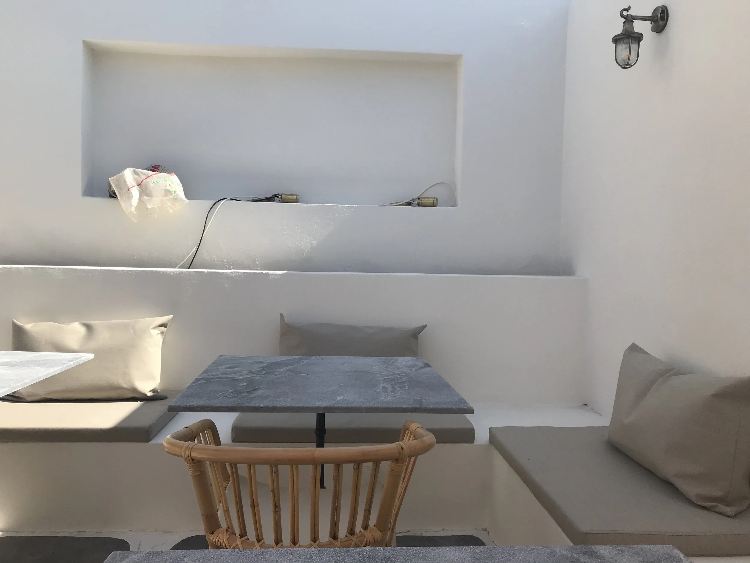 A minimalist interior with white walls and concrete surfaces. There is a wall-mounted lantern on the right, a built-in shelf with objects, a pillow on a ledge, and a wooden chair in the foreground.