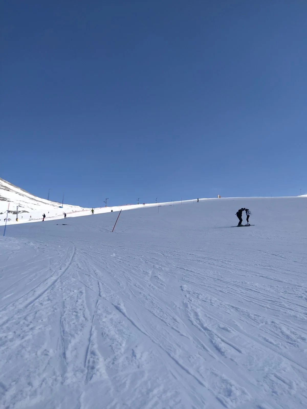 Snow-covered ski slope with skiers and snowboarders under a clear blue sky during daytime.