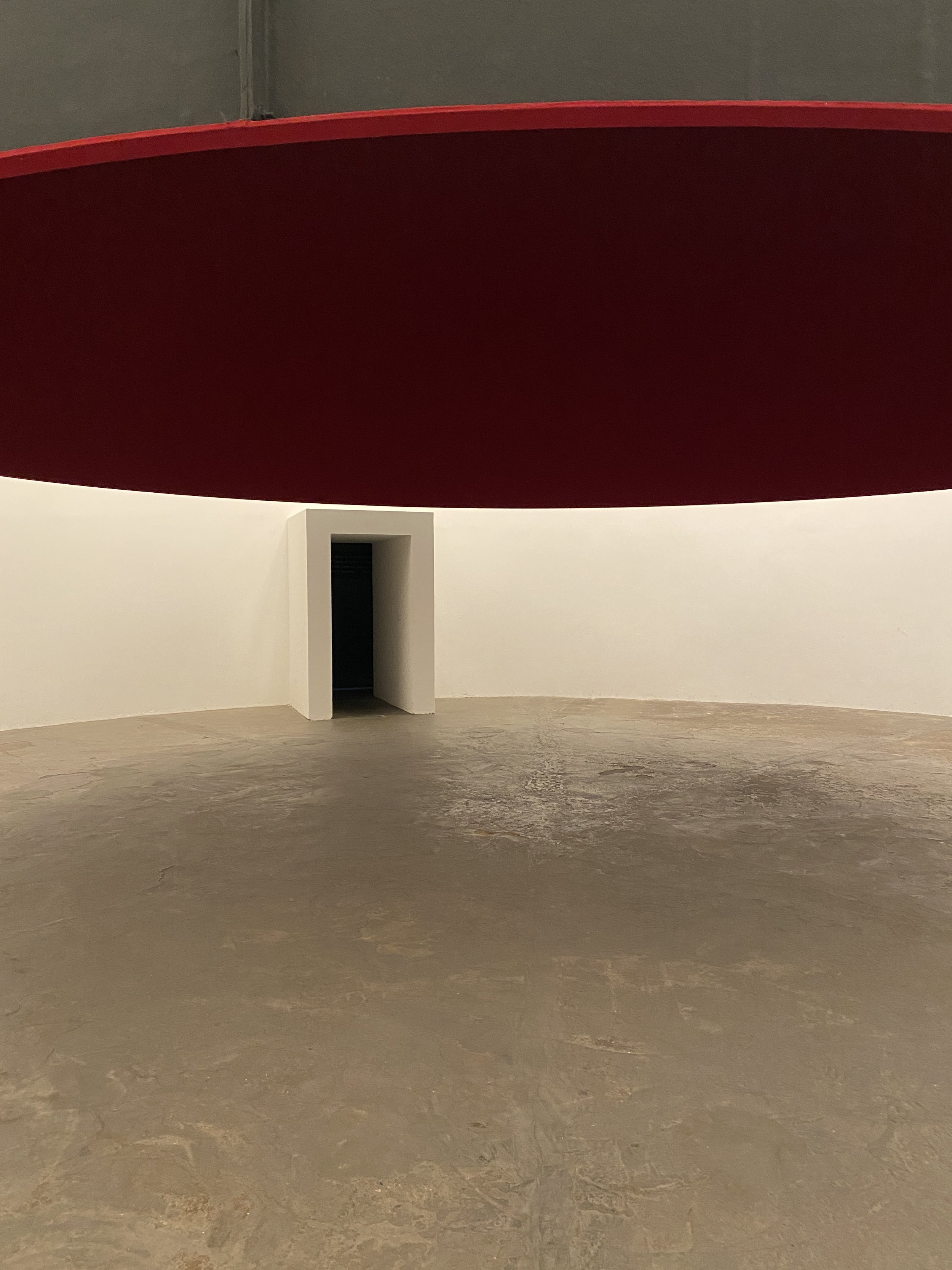 A minimalist interior space featuring a curved white wall with a small rectangular doorway, a smooth beige concrete floor, and a large red curved structure above.